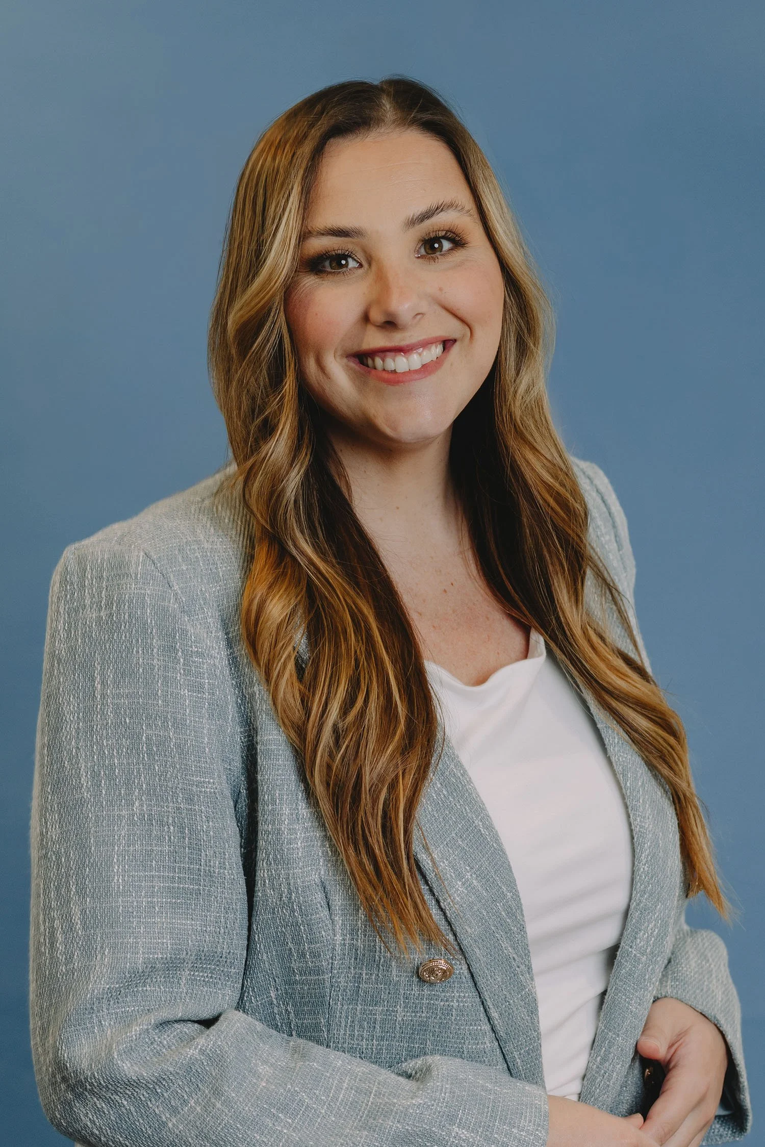 A woman with long wavy brown hair, smiling, wearing a light gray blazer and a white top, standing against a solid blue background.