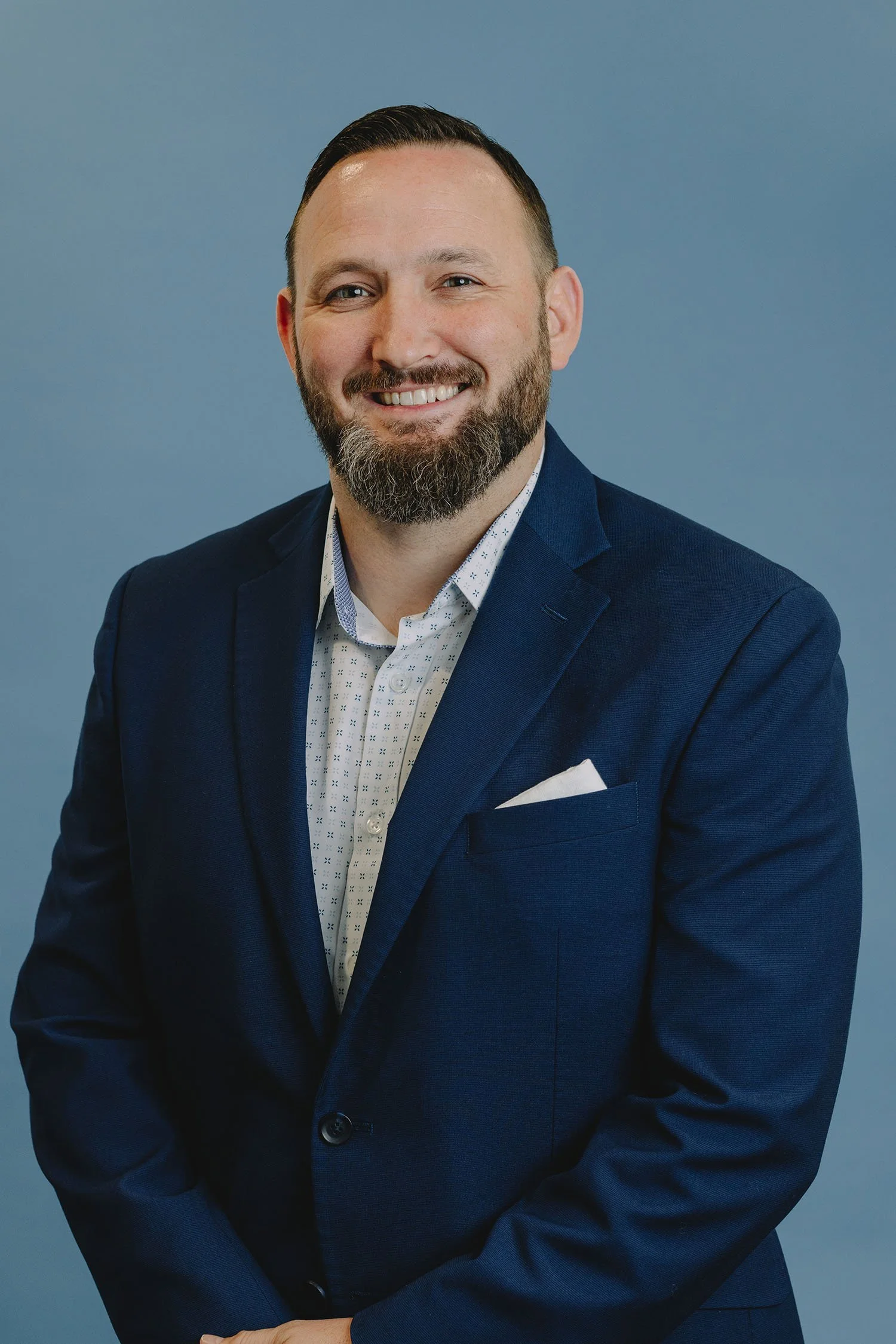 A man with a beard and short hair smiling, dressed in a navy blue suit with a white patterned shirt, standing against a plain blue background.
