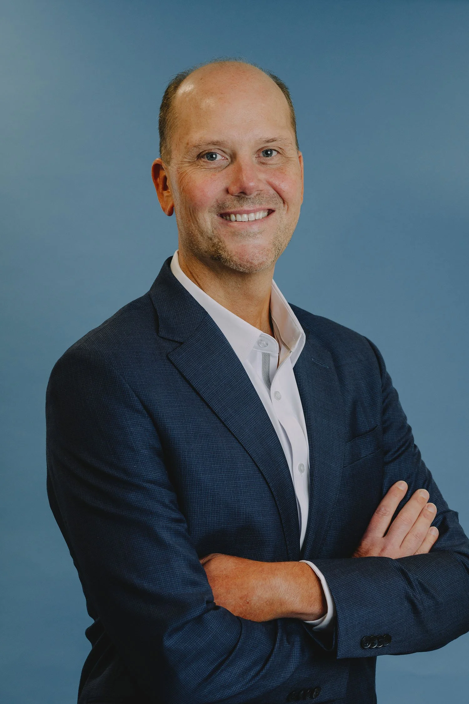 A man in a navy blazer and white shirt, smiling with crossed arms, posing against a plain blue background.