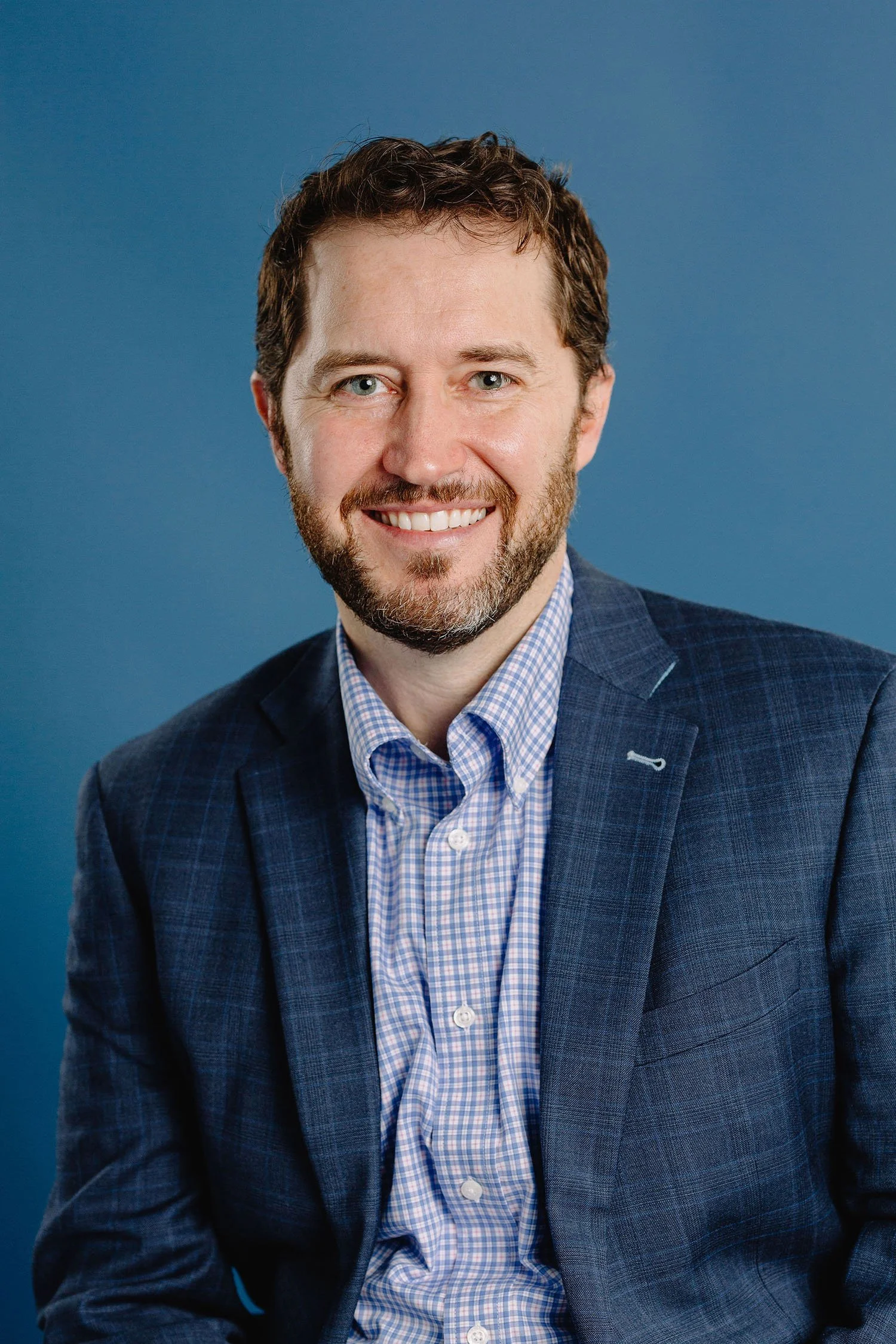 A smiling man with a beard and short curly hair wearing a blue checkered shirt and a dark blue blazer against a plain blue background.