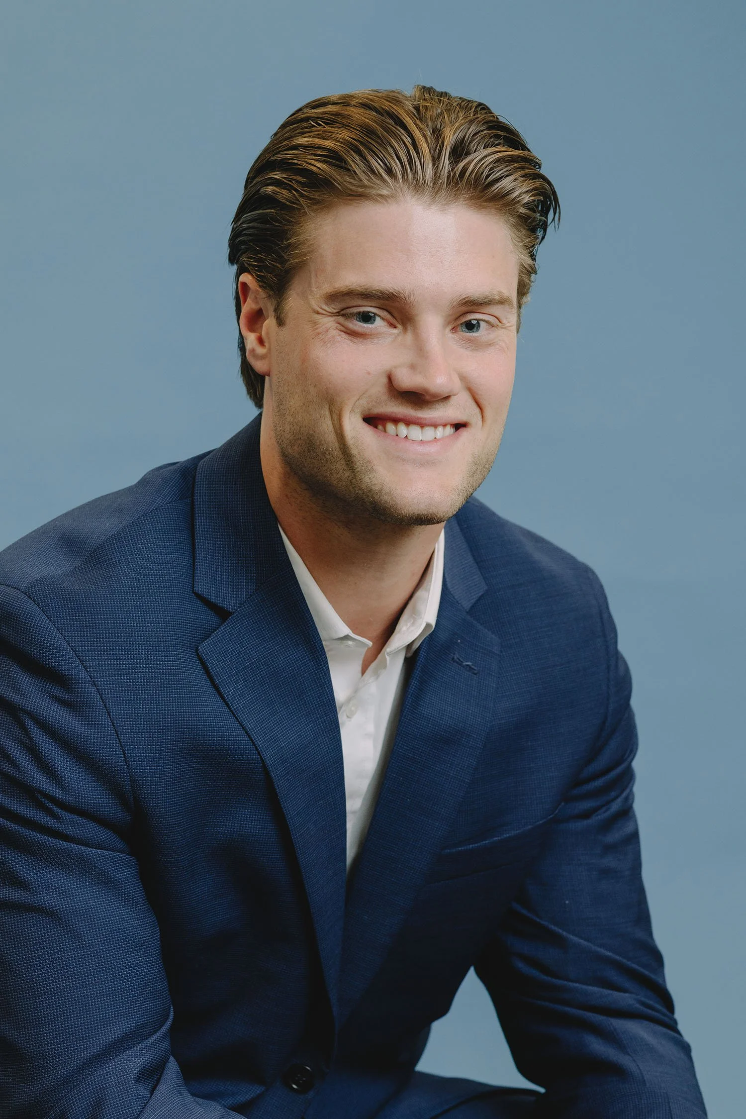 Portrait of a young man with brown hair and blue eyes, wearing a navy blue suit and white shirt, smiling against a plain blue background.