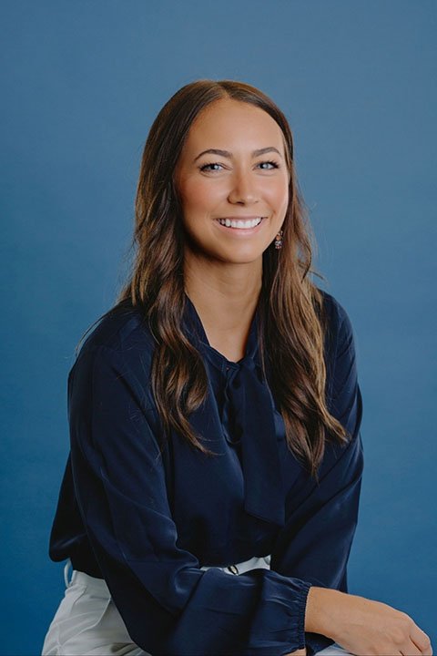 A woman with long brown hair, smiling and wearing a navy blue blouse, sitting against a blue background.
