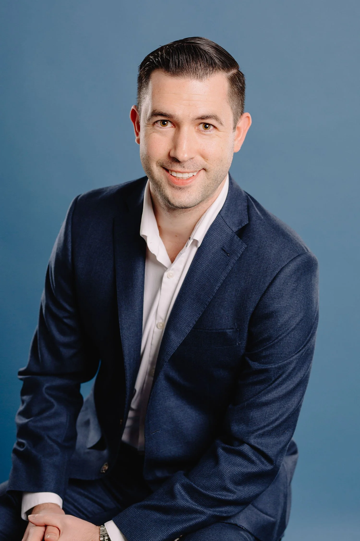 Portrait of a man with dark, slicked-back hair, wearing a dark blue suit and white shirt, seated against a blue background, smiling slightly.