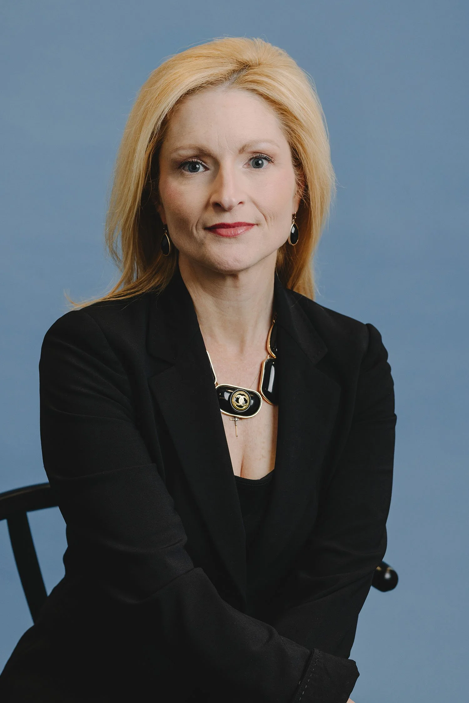 A woman with blonde hair and blue eyes wearing a black blazer and jewelry, sitting in front of a blue background.