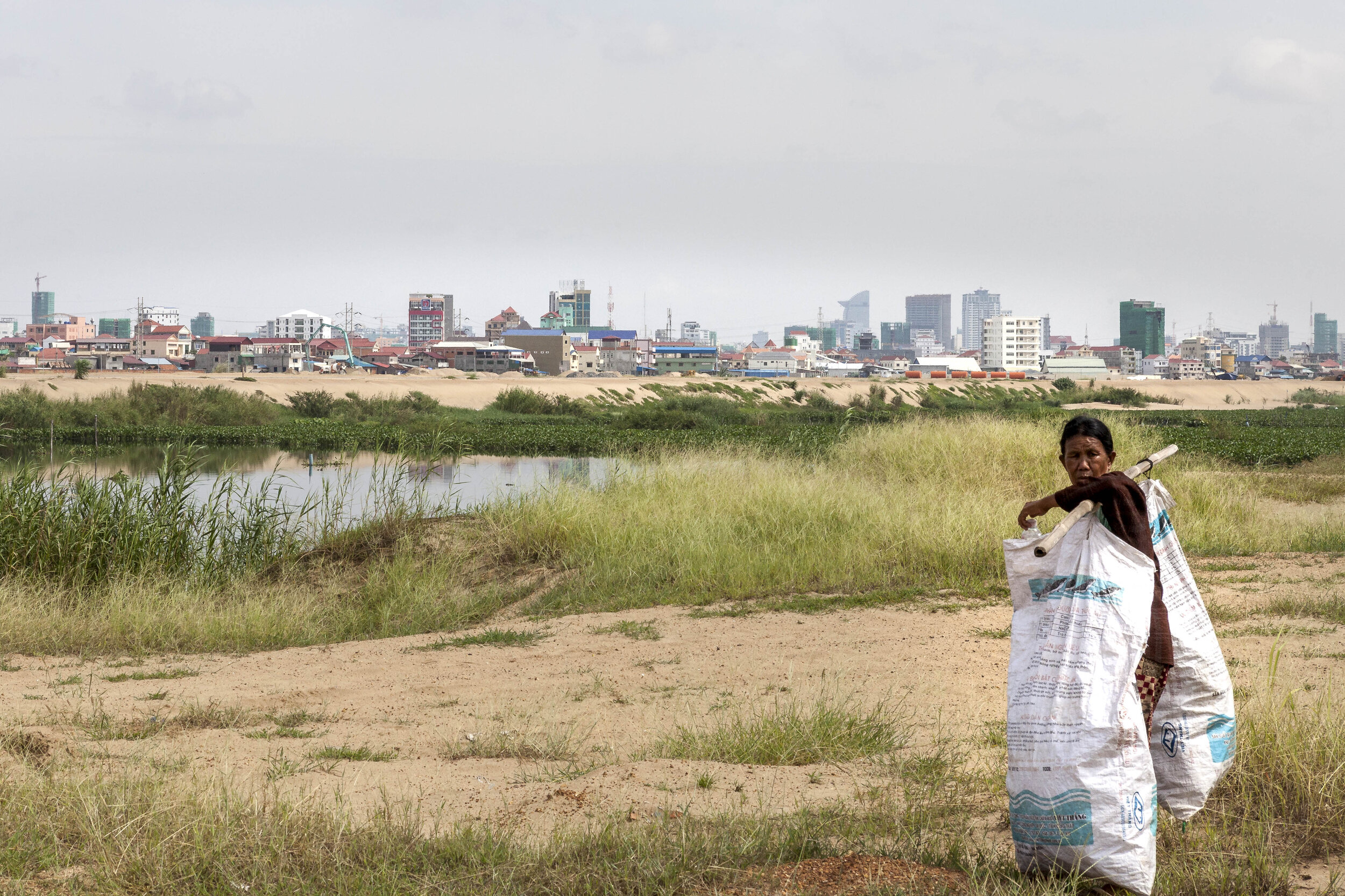  A woman collects plastic rubbish, with the Phnom Penh skyline and a filled portion of the lake behind her. 