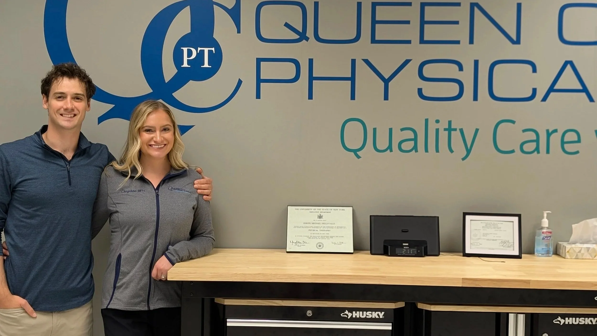 Two smiling people, a man and a woman, standing next to each other at a reception desk in a medical or physical therapy office. The background shows a wall with the logo and name of the facility, 'Queen City Physical Therapy,' and some framed documen