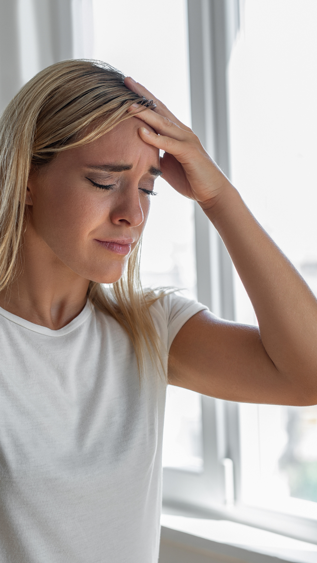 Woman in white shirt holding her head in pain, appearing to have a headache, standing by a window.