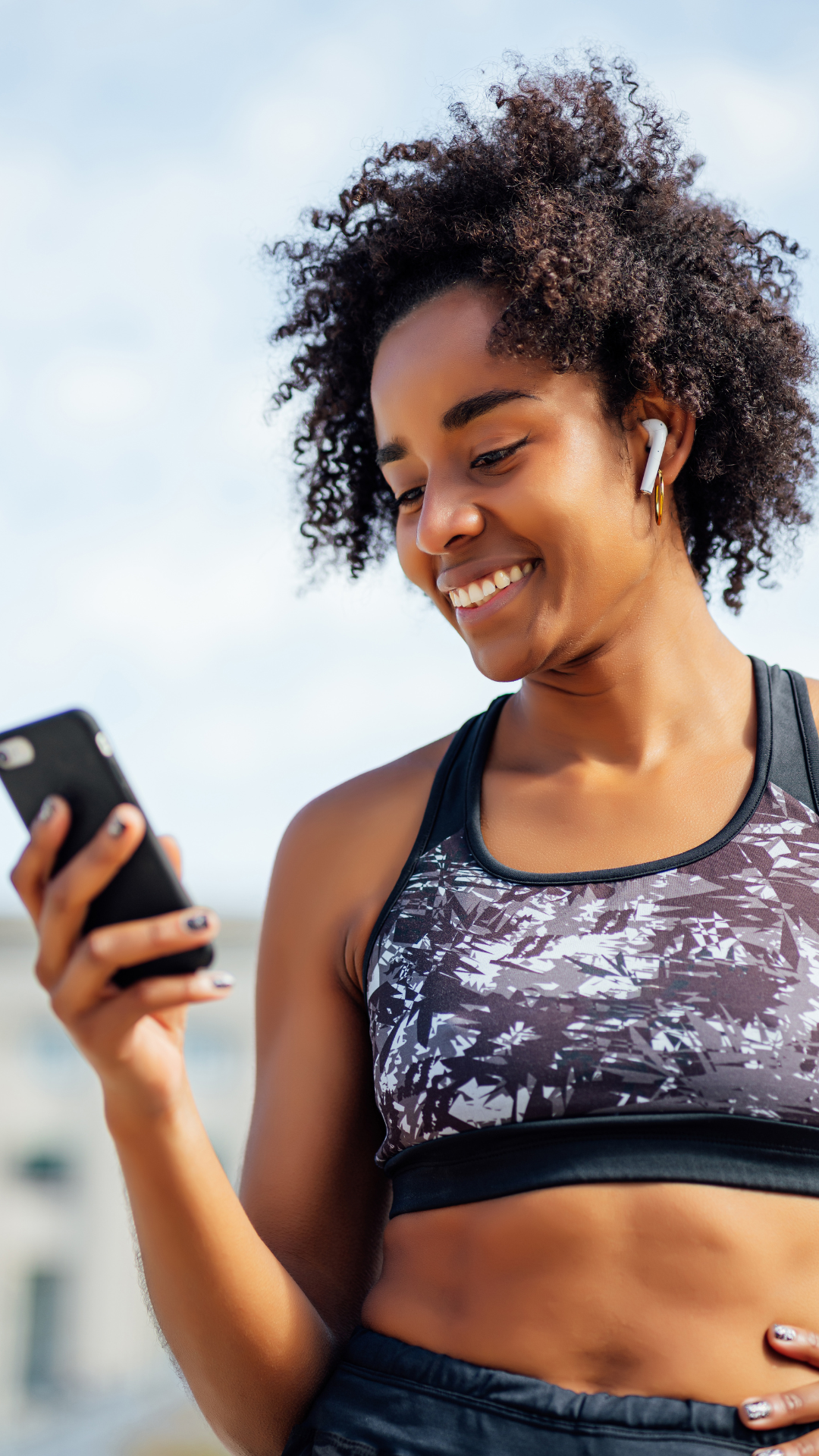 Woman in workout clothes smiling while looking at smartphone and wearing wireless earbuds.