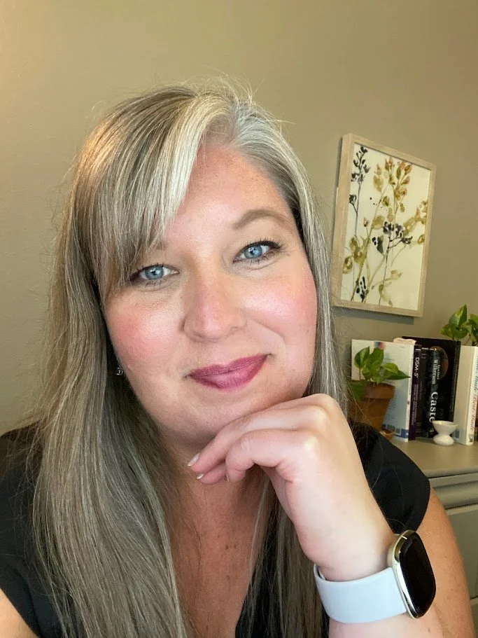 A woman with long gray hair and blue eyes, wearing a black shirt and a wristwatch, resting her chin on her hand in an office with books and a framed botanical print.