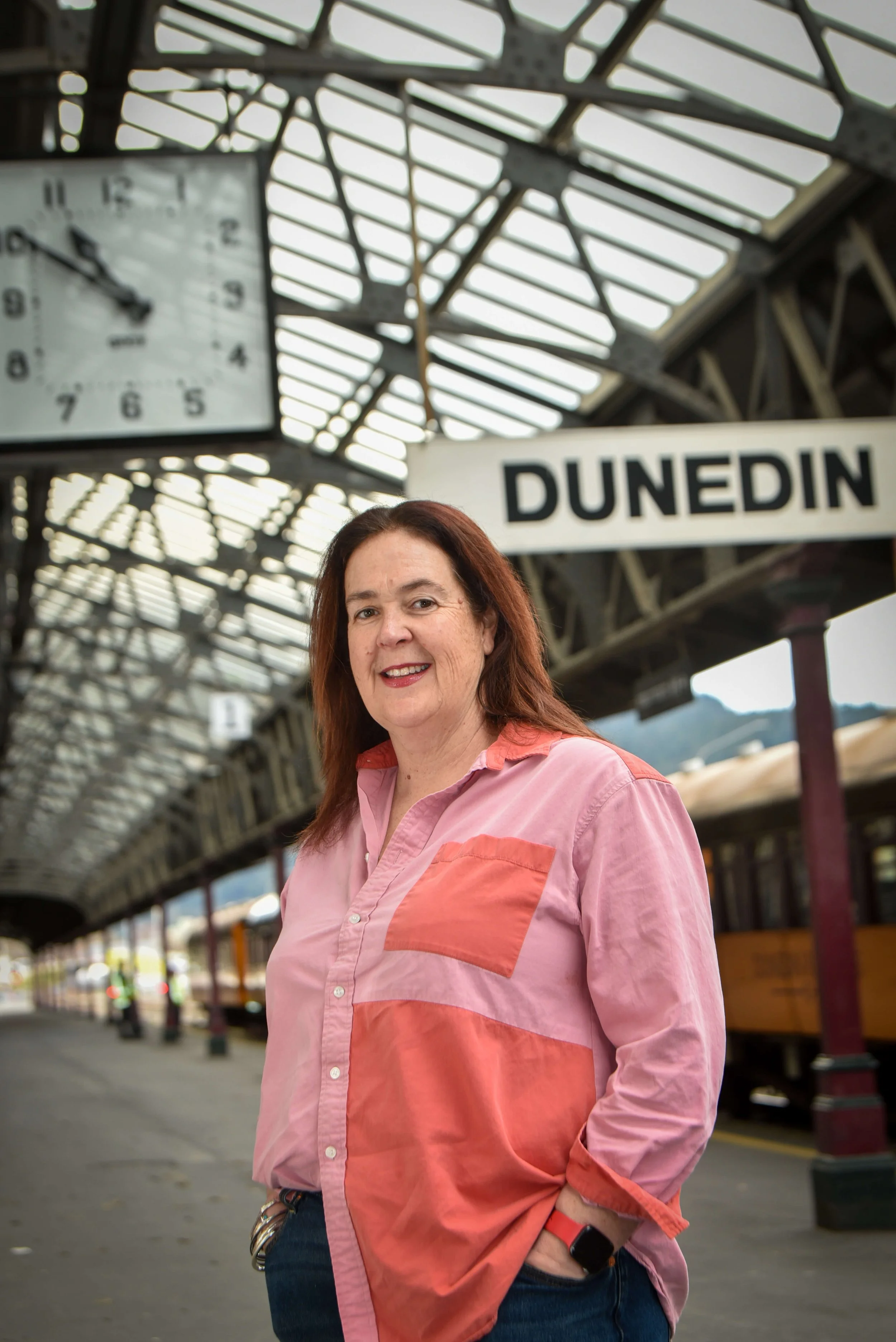 A woman with red hair smiling at a train station in Dunedin, wearing a pink and orange shirt and jeans, with a watch on her left wrist and keys clipped to her pocket.