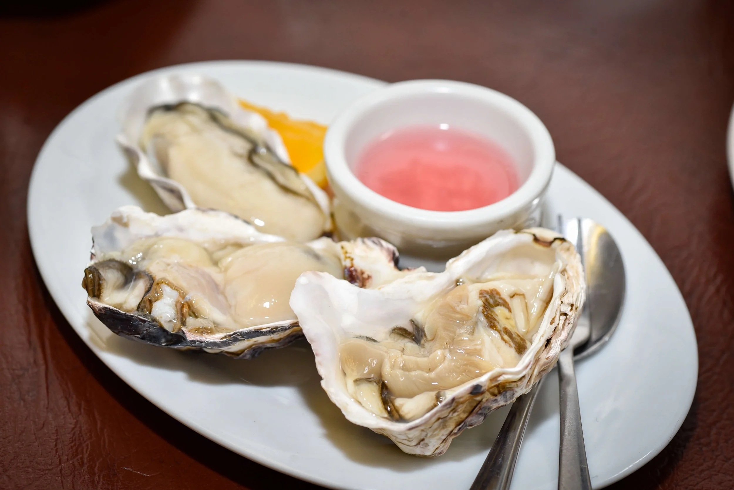 Fresh oysters on a white plate with a small bowl of pink sauce and a silver utensil, on a dark wooden table.