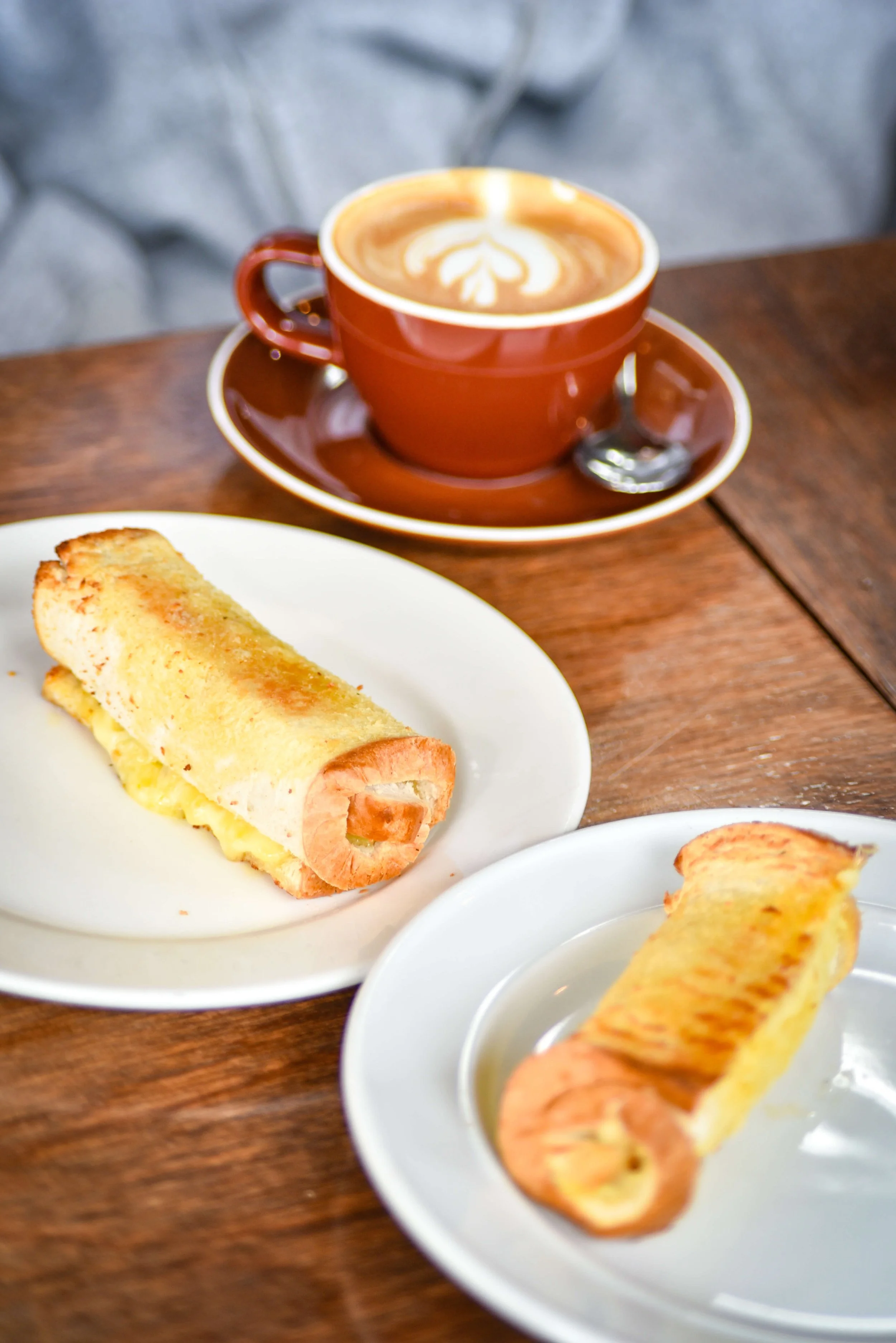 A cup of coffee with latte art on a saucer, and two breakfast wraps on separate plates on a wooden table.