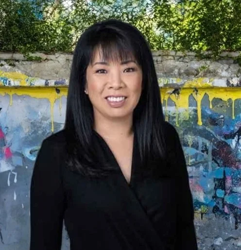 Coreen standing in front of a graffiti-covered wall with yellow accents and green trees in the background.