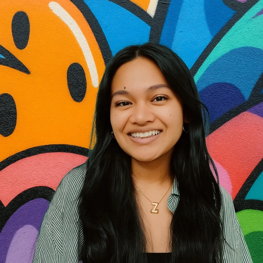 A smiling young woman with long black hair, wearing a striped shirt and a gold necklace with a 'Z', standing against a colorful graffiti wall.