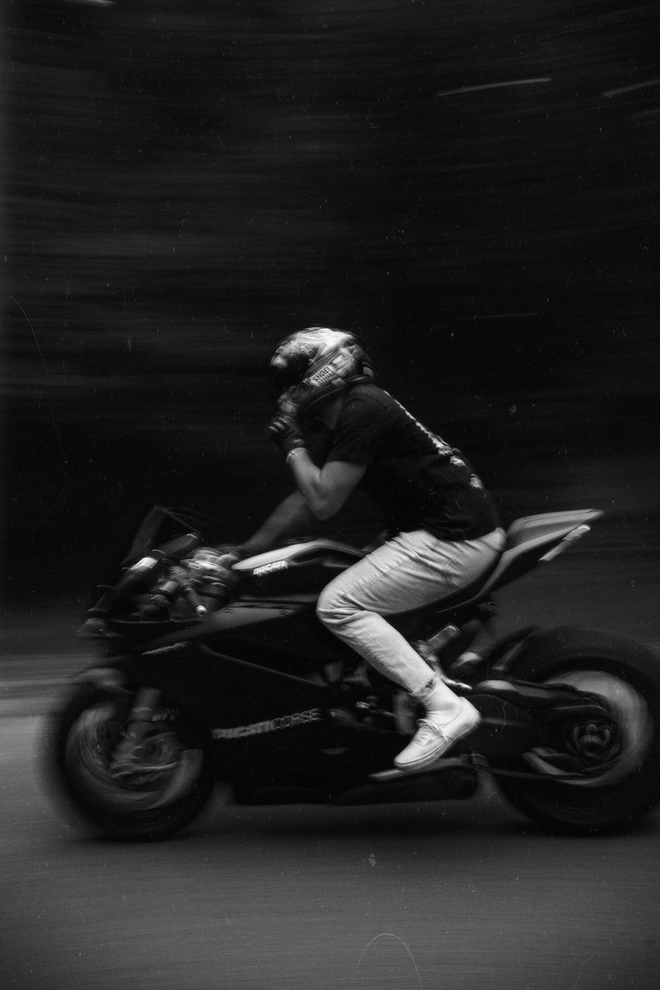 Person riding a motorcycle at high speed, wearing a helmet and casual clothing, in a blurred motion background.