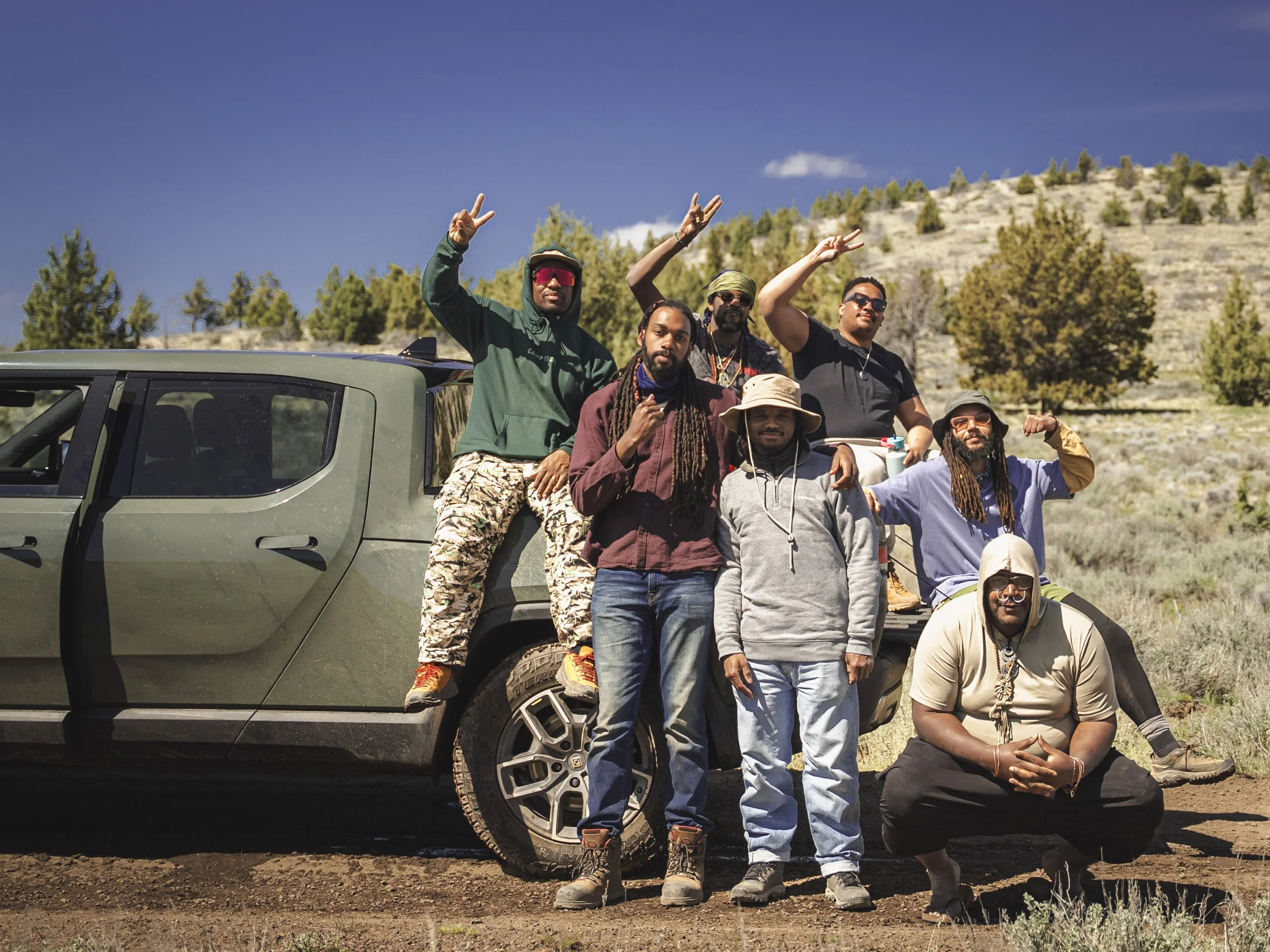 Seven Black men are posed in front of a truck in a dessert backdrop. Some men are holding up a peace sign with their hands.