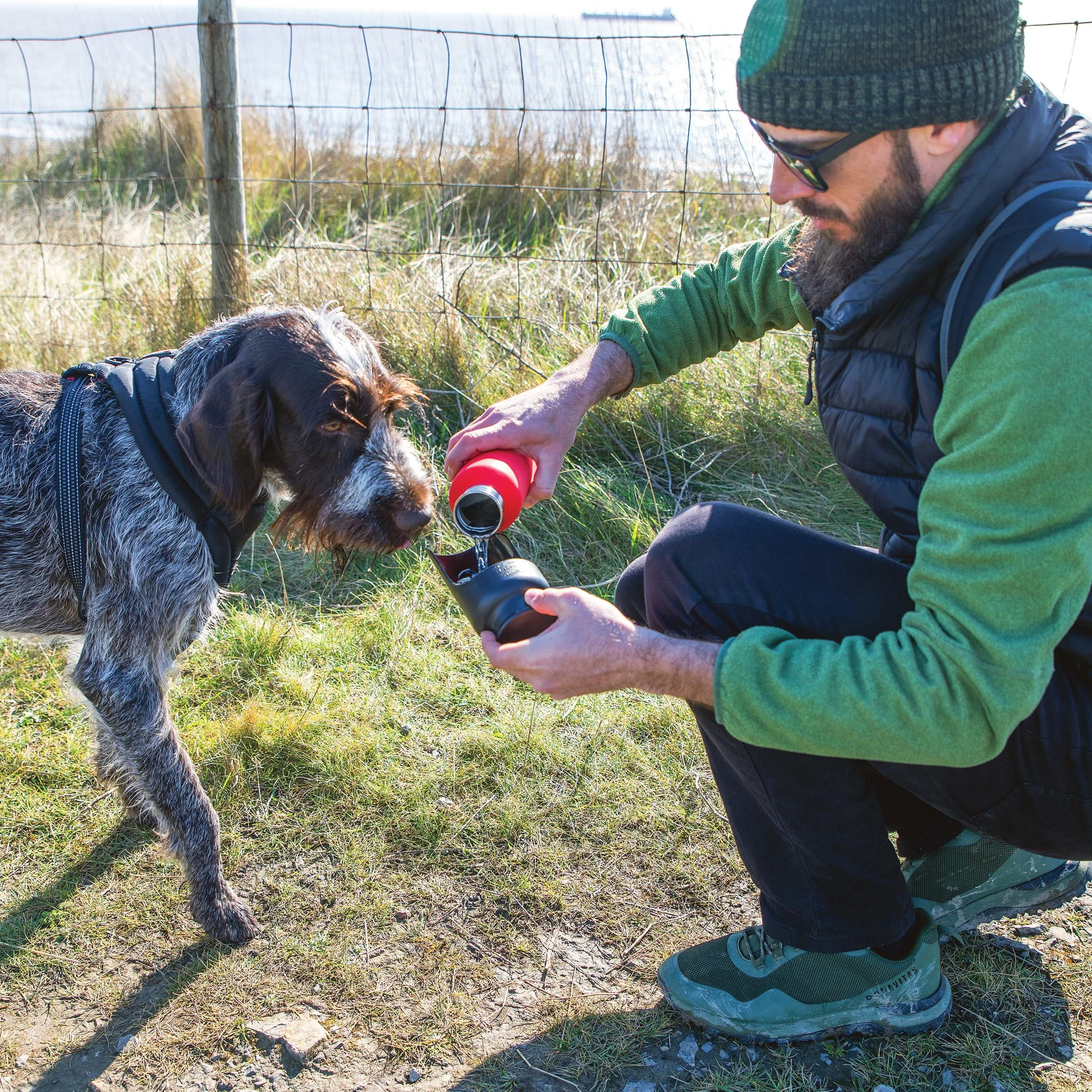 A man in outdoor clothing kneels to give water from a bottle to a dog outdoors near a fence.