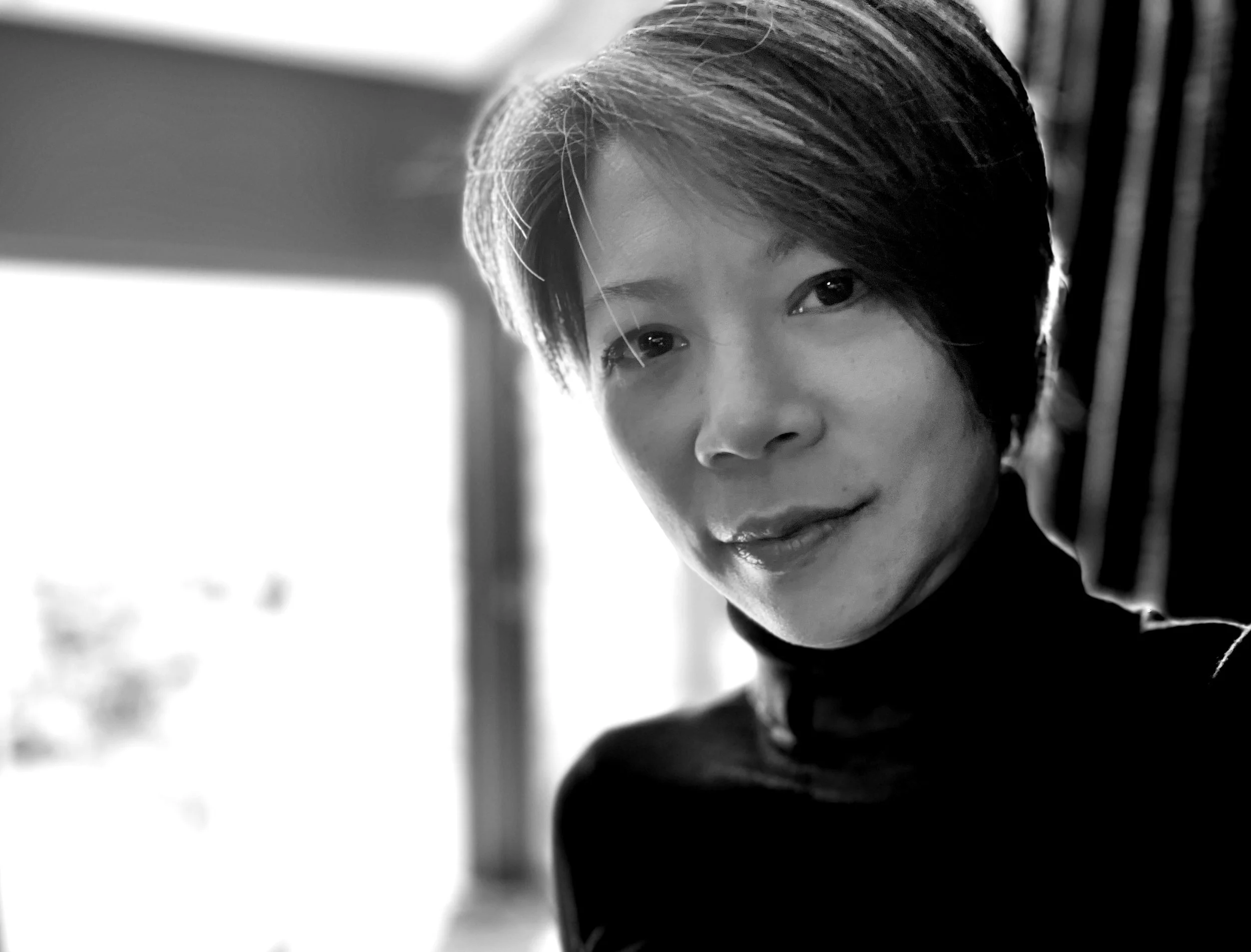 Black and white portrait of a woman with short hair, smiling softly, wearing a turtleneck, in front of a bright background.