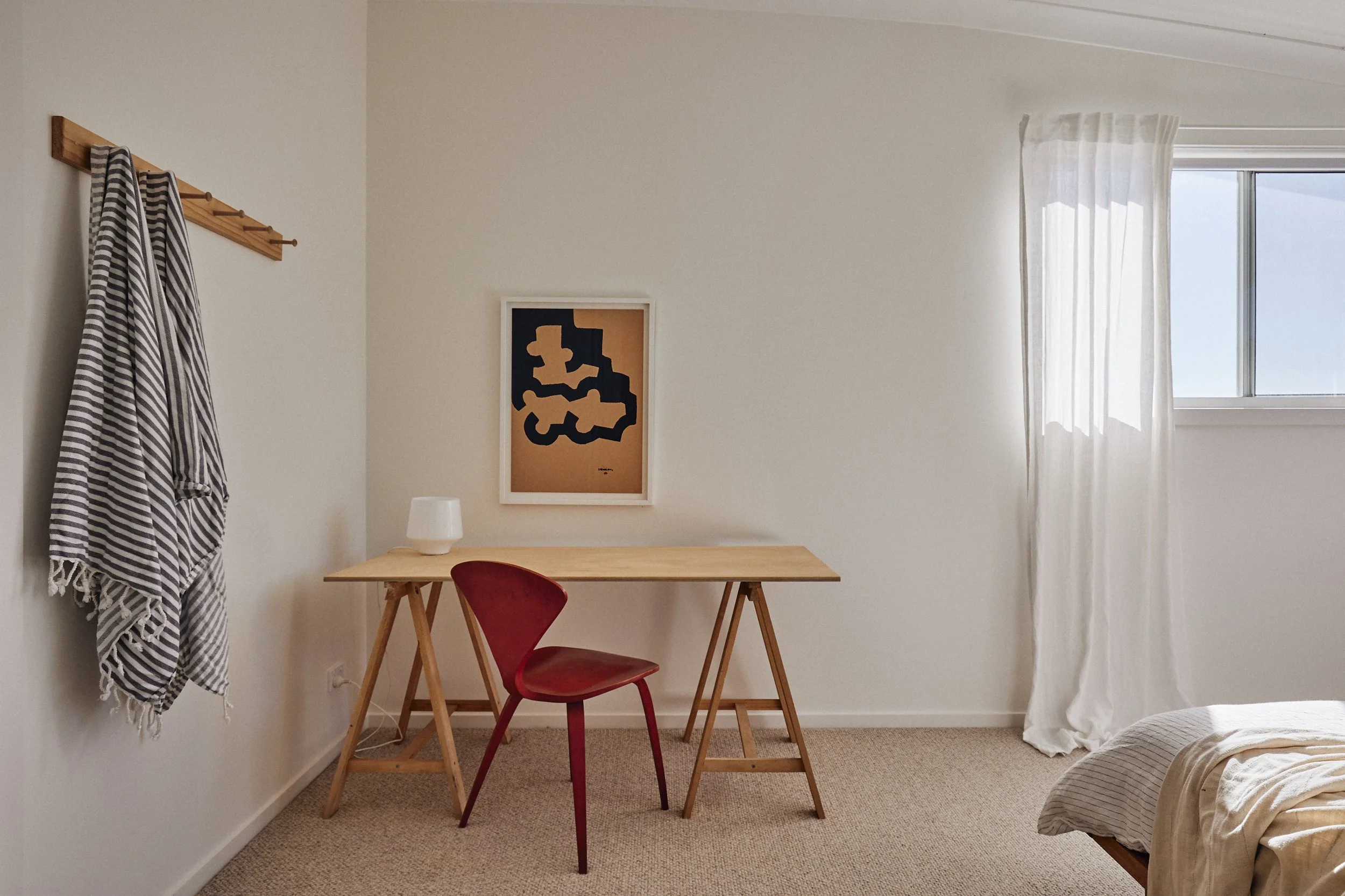 A minimalist bedroom with a wooden desk, a red chair, a white lamp, and a framed abstract artwork on the wall. There are white curtains on the window, and part of a bed is visible with beige linens. A striped cloth hangs on a wooden hook on the wall.