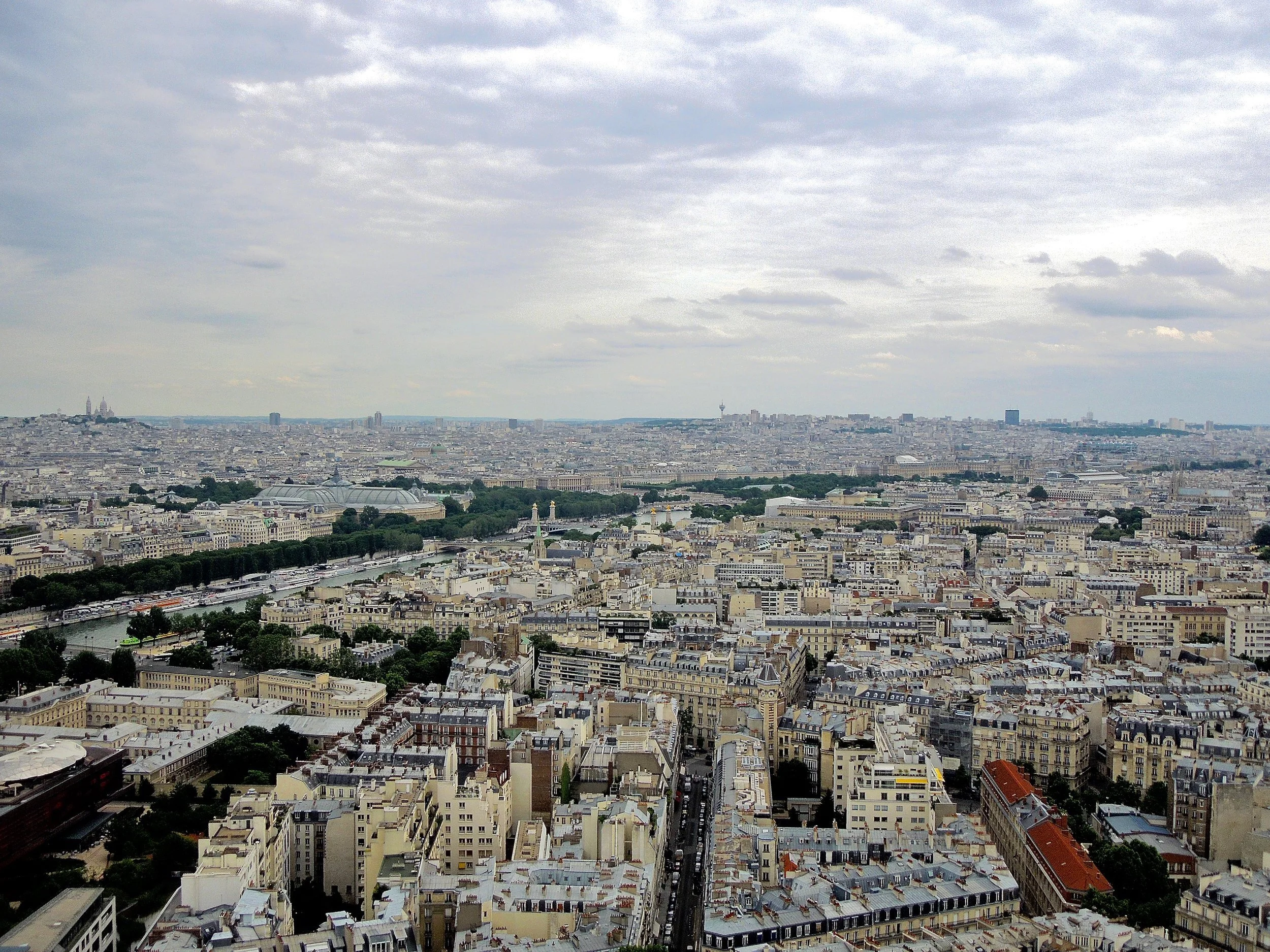 Panoramic view of Paris, France, featuring a dense urban landscape with numerous historical buildings, the River Seine, and overcast skies.