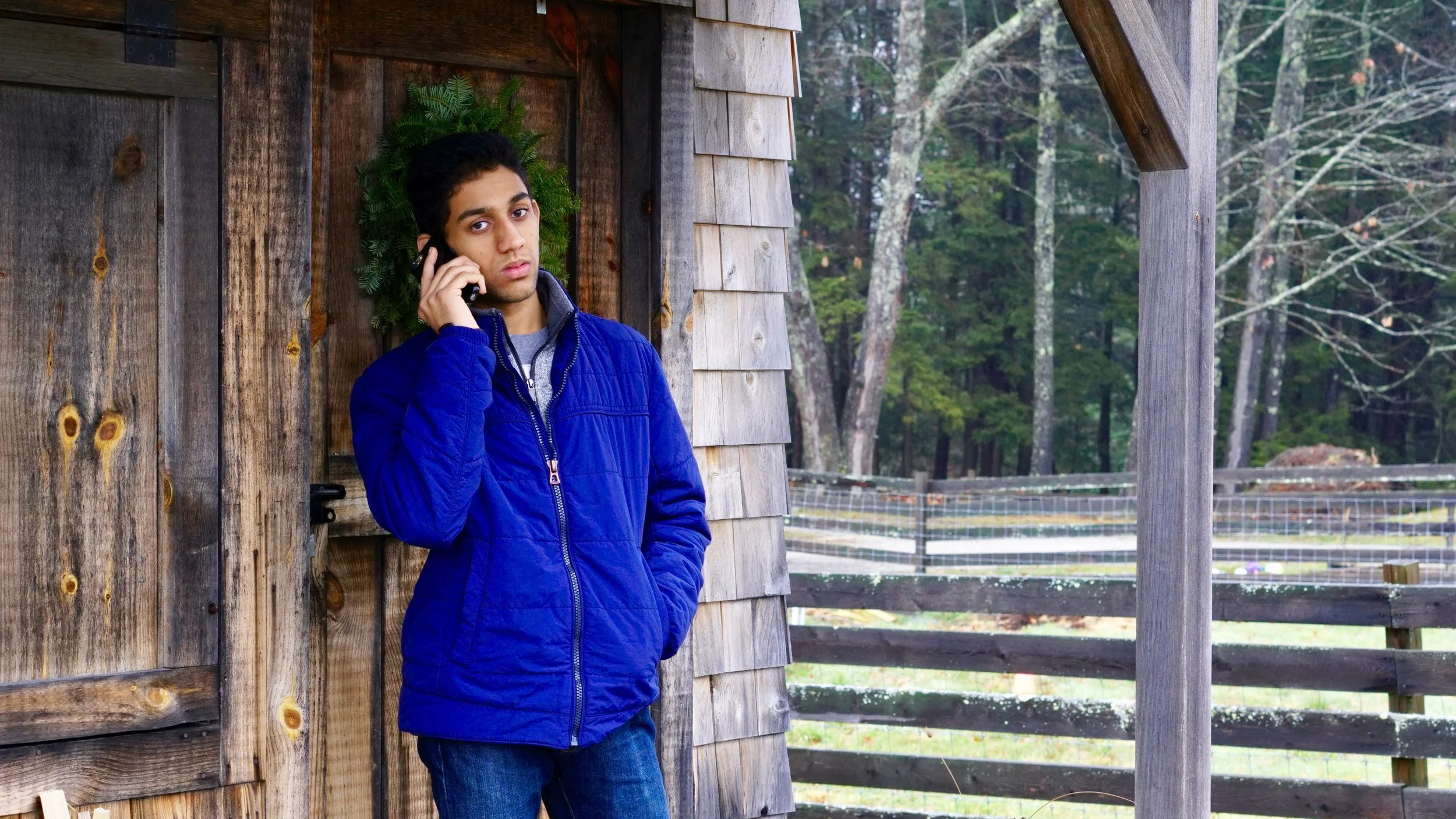 Person wearing a blue jacket talking on a cellphone, leaning against a wooden cabin with a forest in the background.