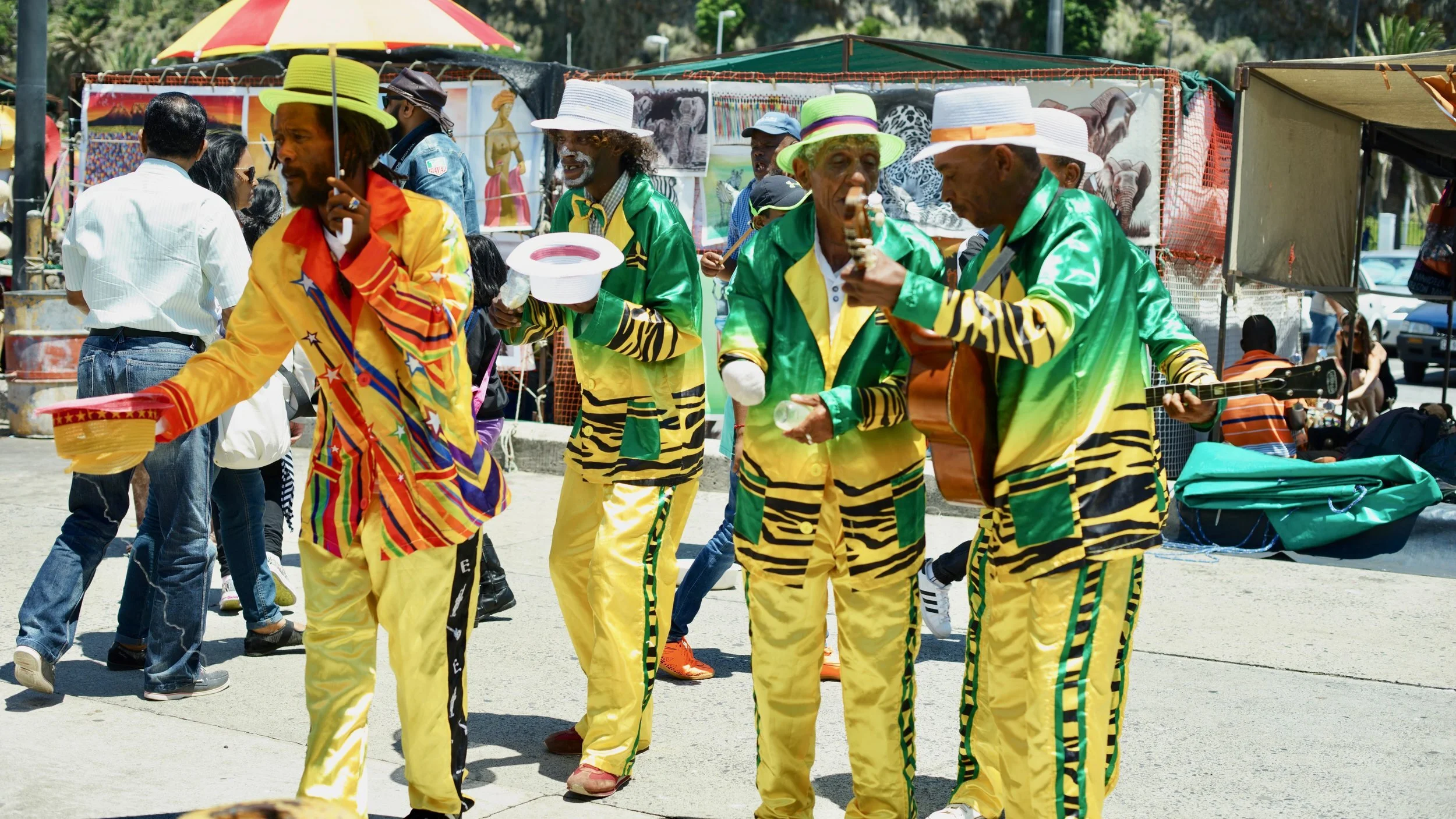 Street performers in colorful suits dancing and playing musical instruments in an outdoor market setting, surrounded by people and vendor stalls.