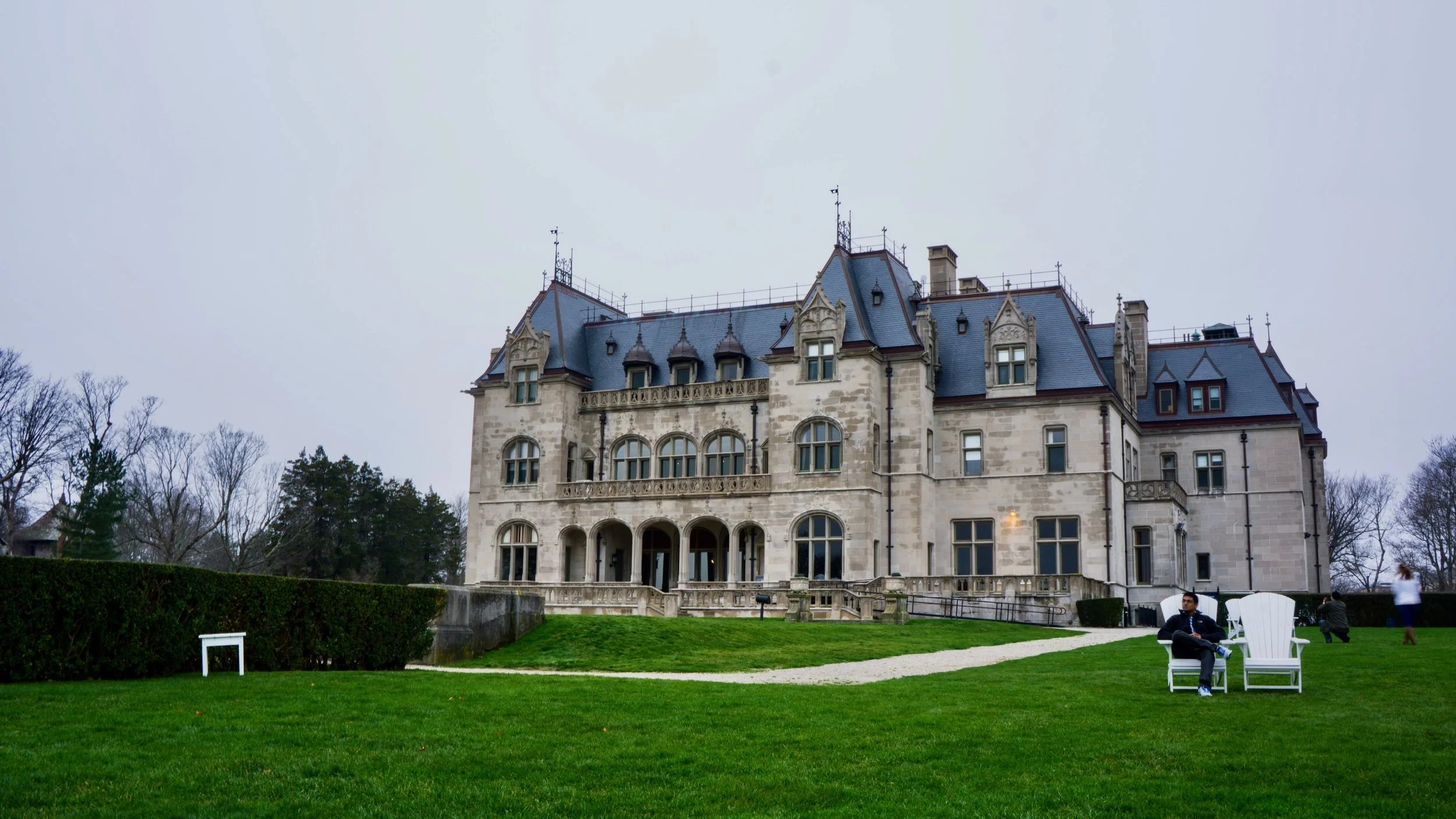 Victorian-style stone mansion with arched windows and decorative gables, surrounded by green lawn and garden benches.