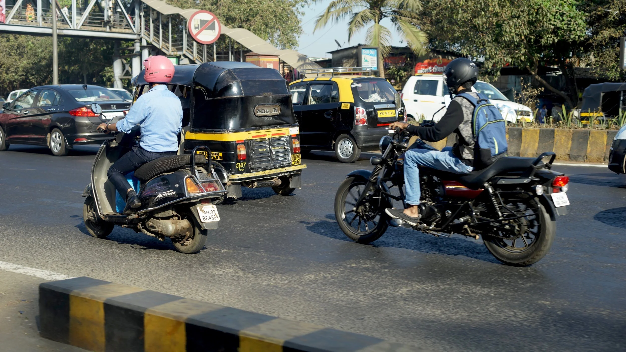 Traffic scene with vehicles and two-wheeler riders, including scooters and motorcycles, on a busy urban road during the day.