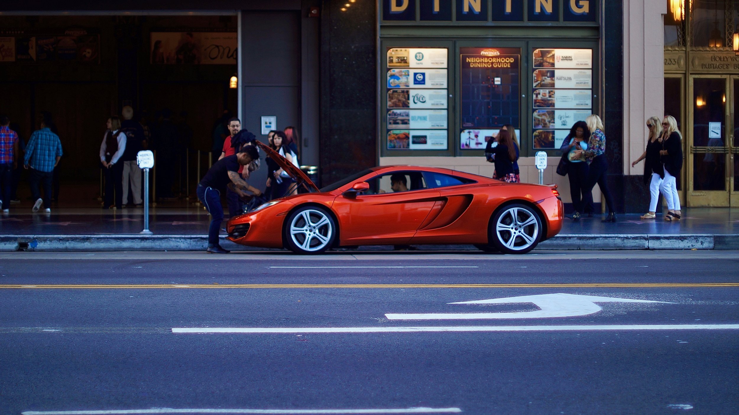 Orange sports car parked on a city street with people nearby, storefronts in the background.