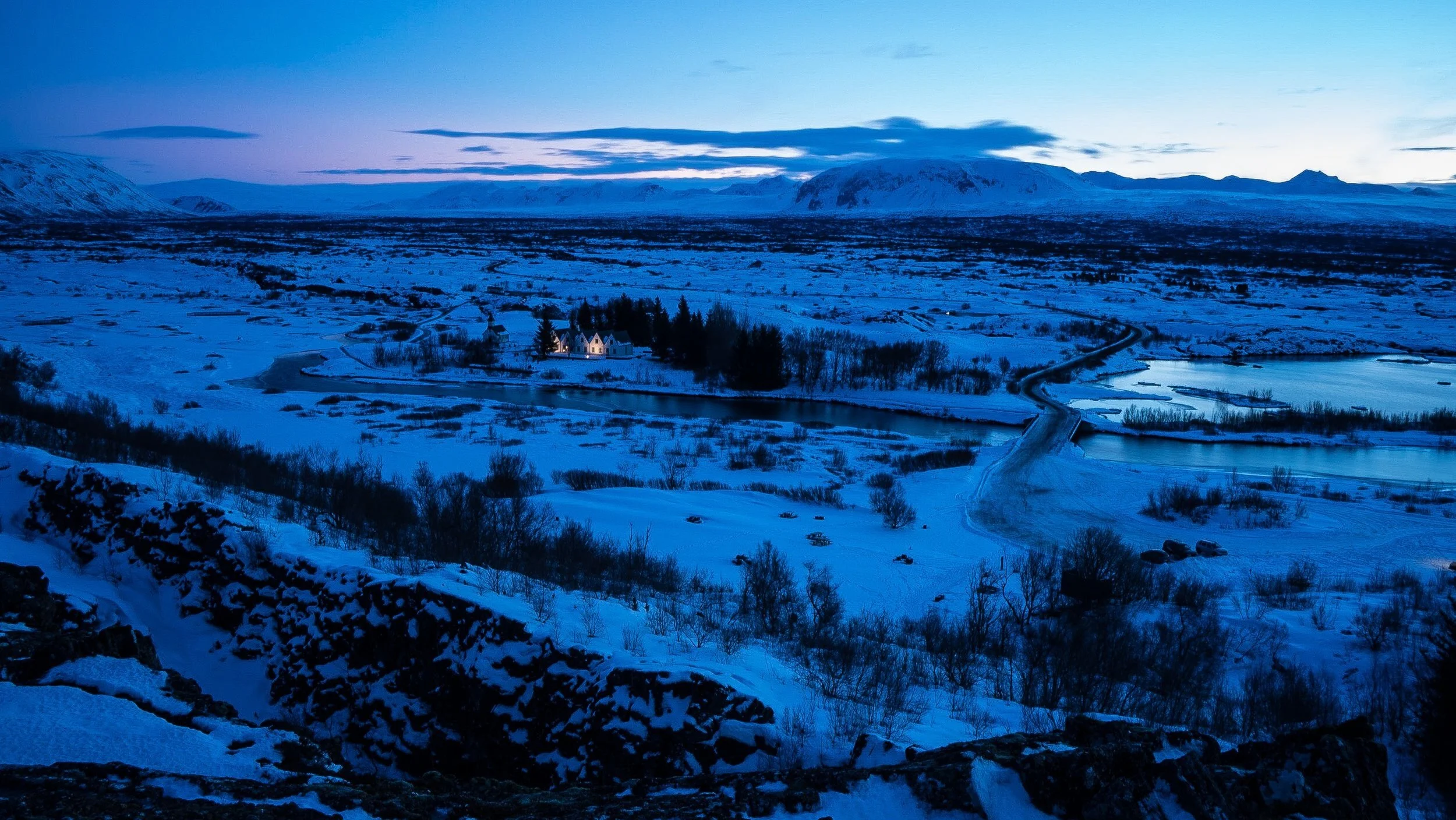 Snow-covered landscape during twilight with a river and distant hills, featuring a small cluster of illuminated buildings surrounded by trees.