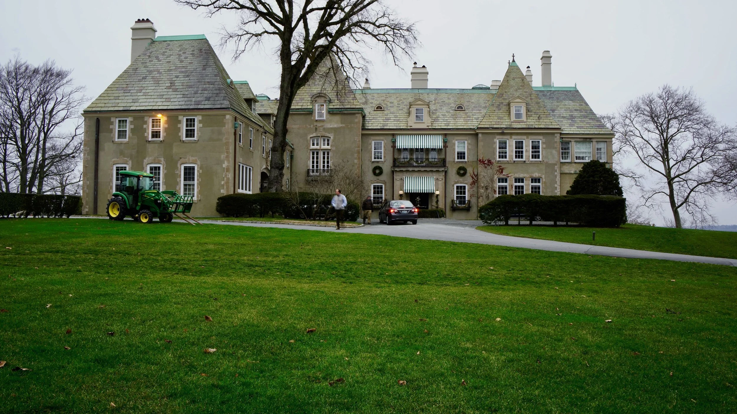 Large historic mansion with a green, manicured lawn, a tractor parked nearby, a car on the driveway, and a person approaching the house; overcast sky.