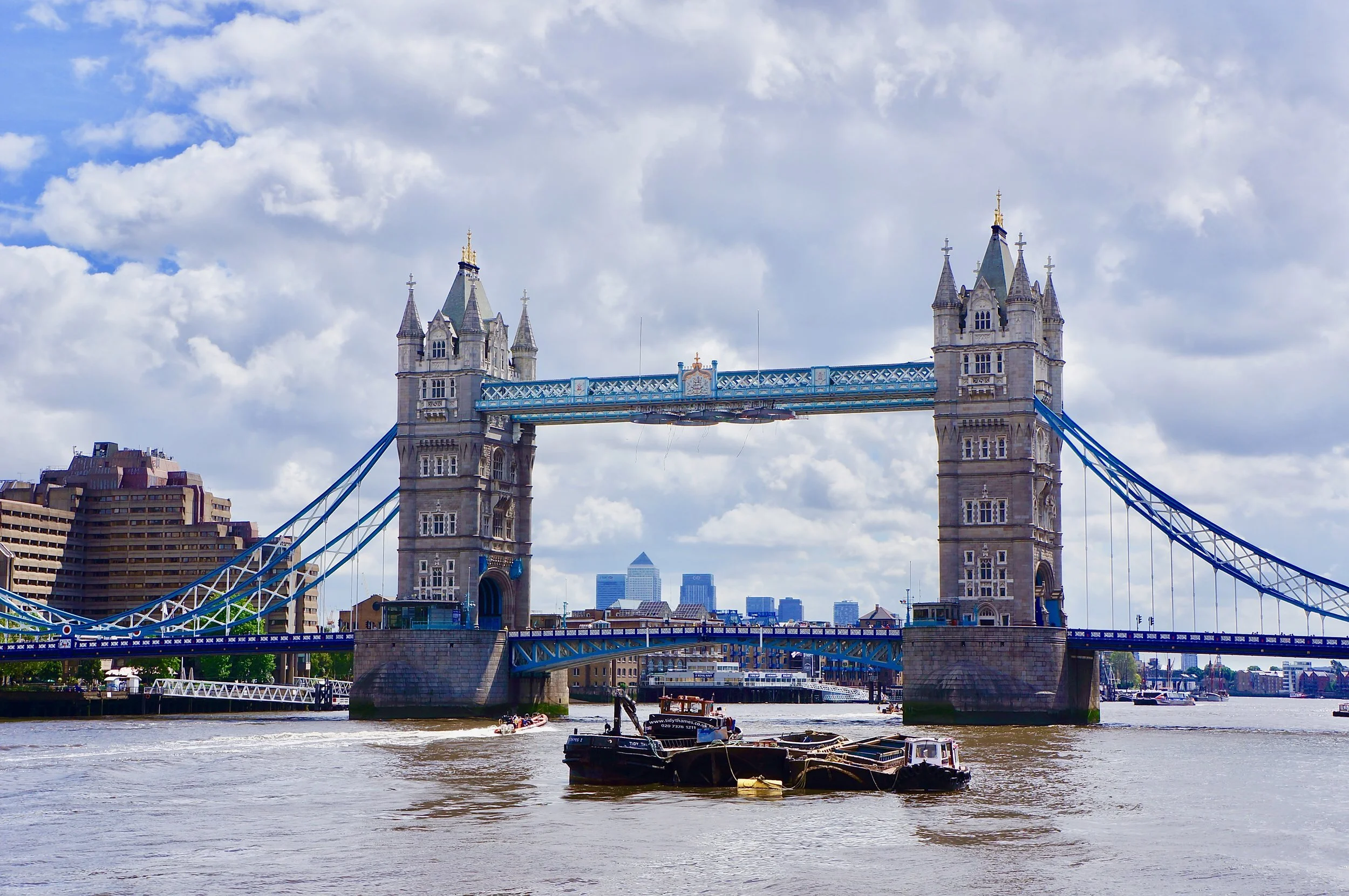 Tower Bridge in London over the River Thames with boats passing underneath and buildings in the background on a cloudy day.
