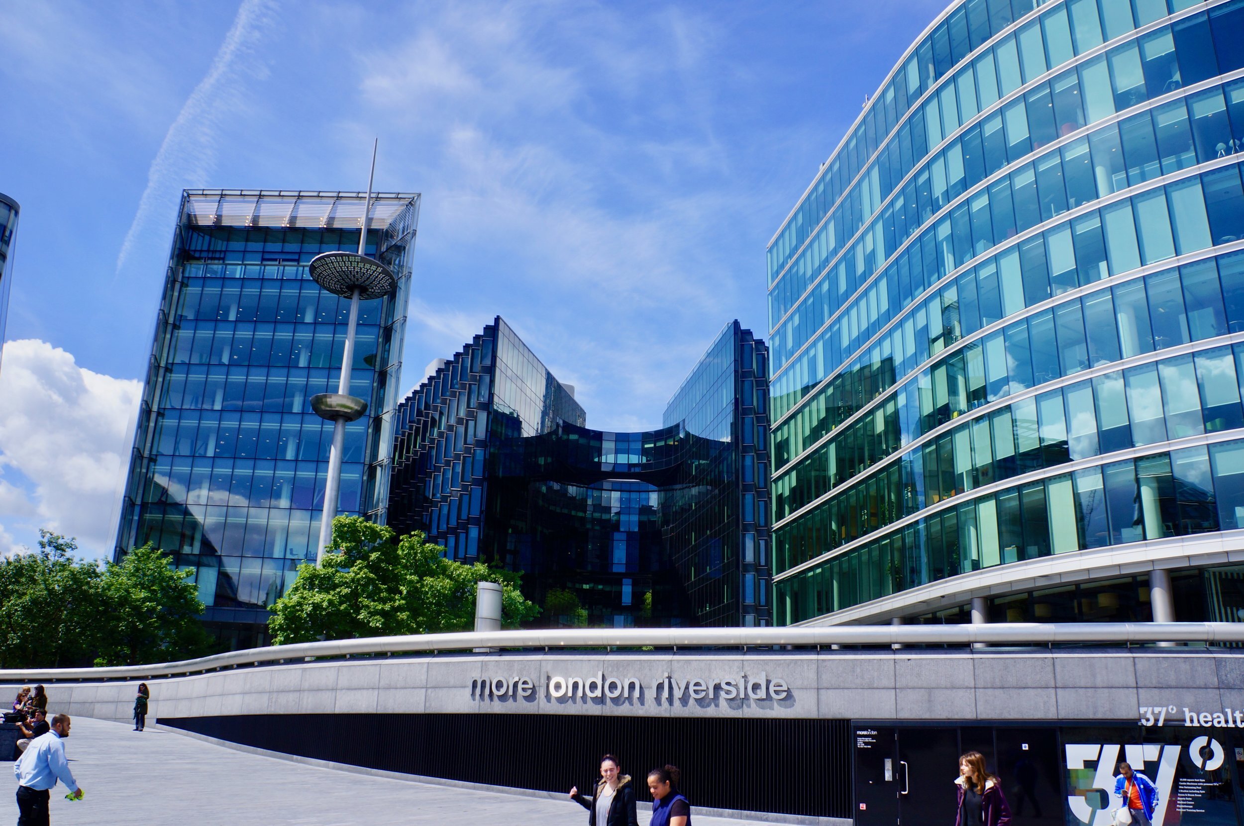 Modern glass office buildings at More London Riverside, sunny day with blue sky, pedestrians walking.