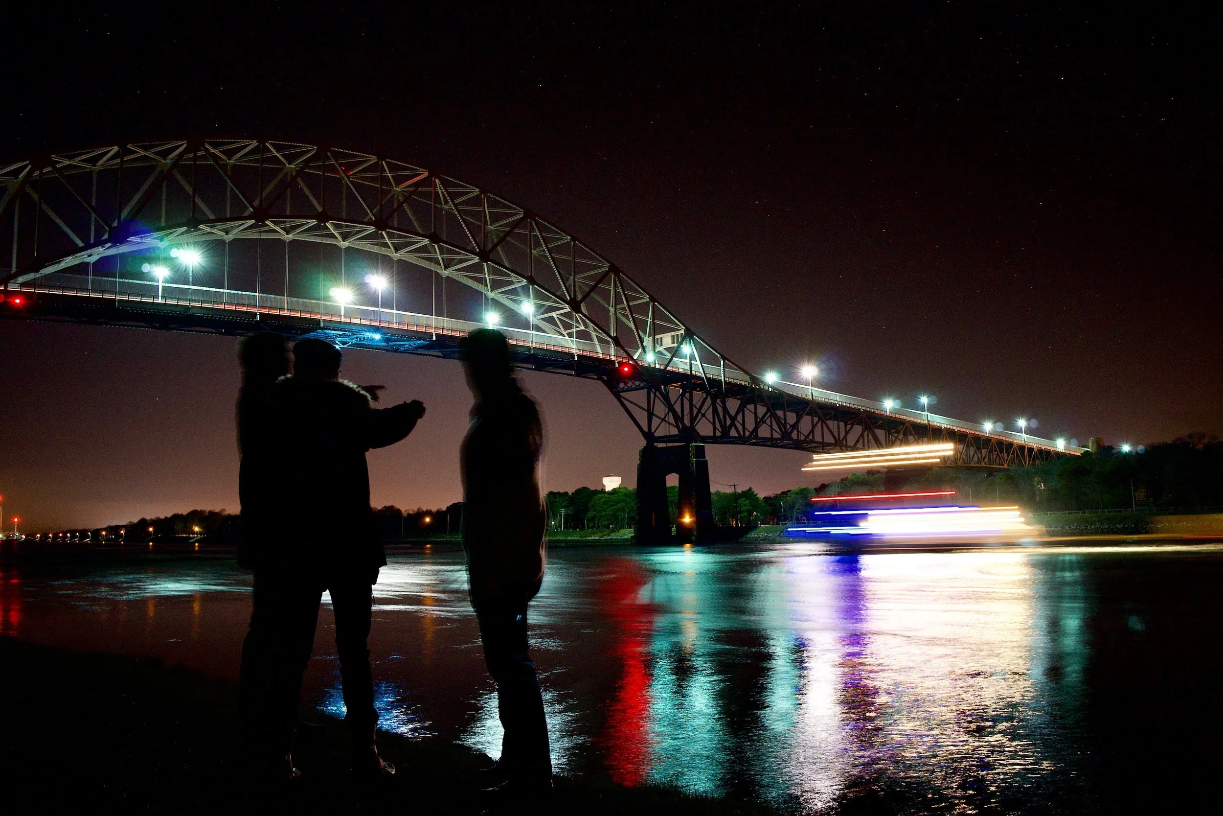 Night scene of a lit bridge with three silhouettes and a boat in motion blur on the water.