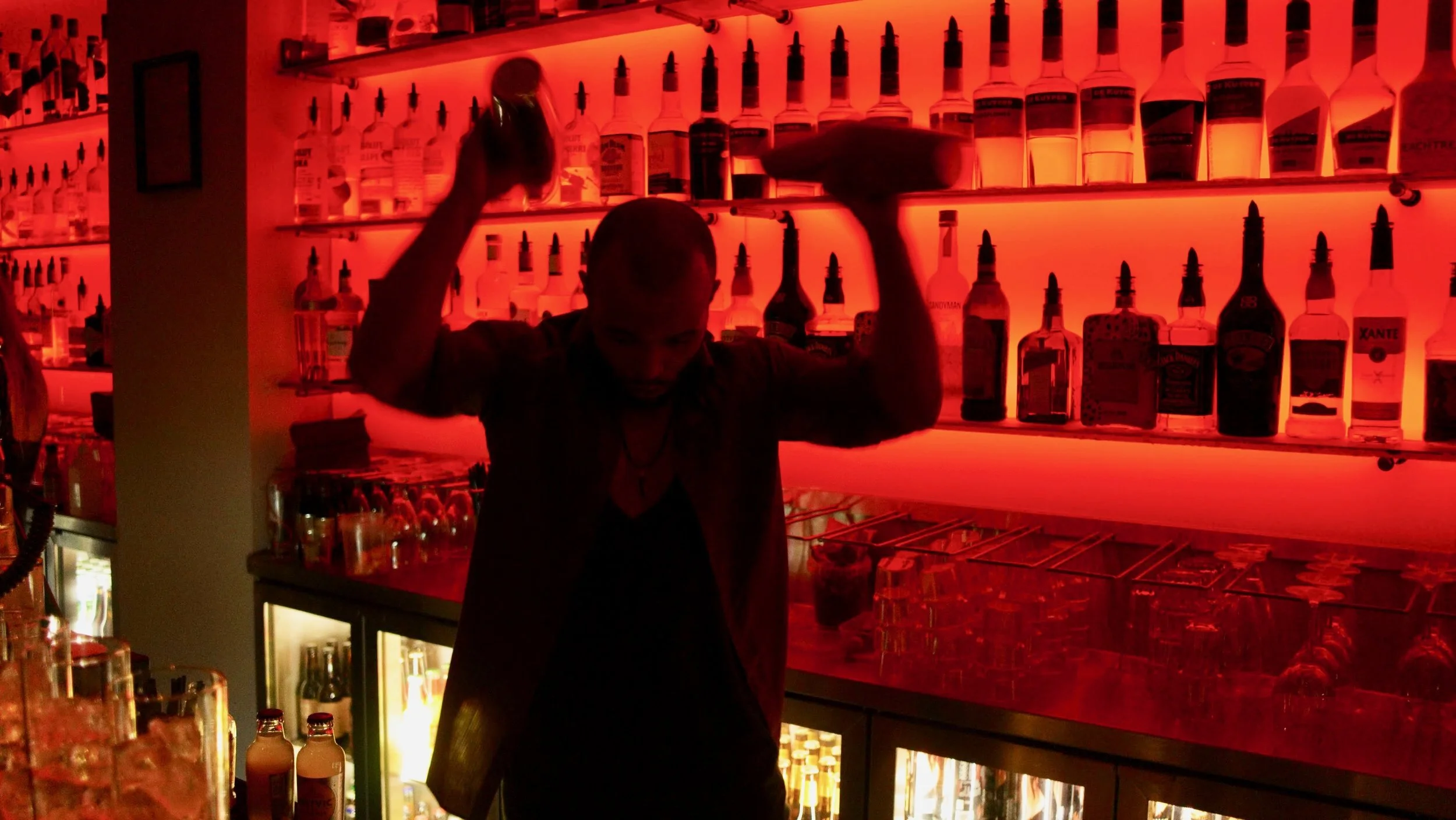 Bartender mixing drinks at a dimly lit bar with red lighting and shelves of alcohol bottles.