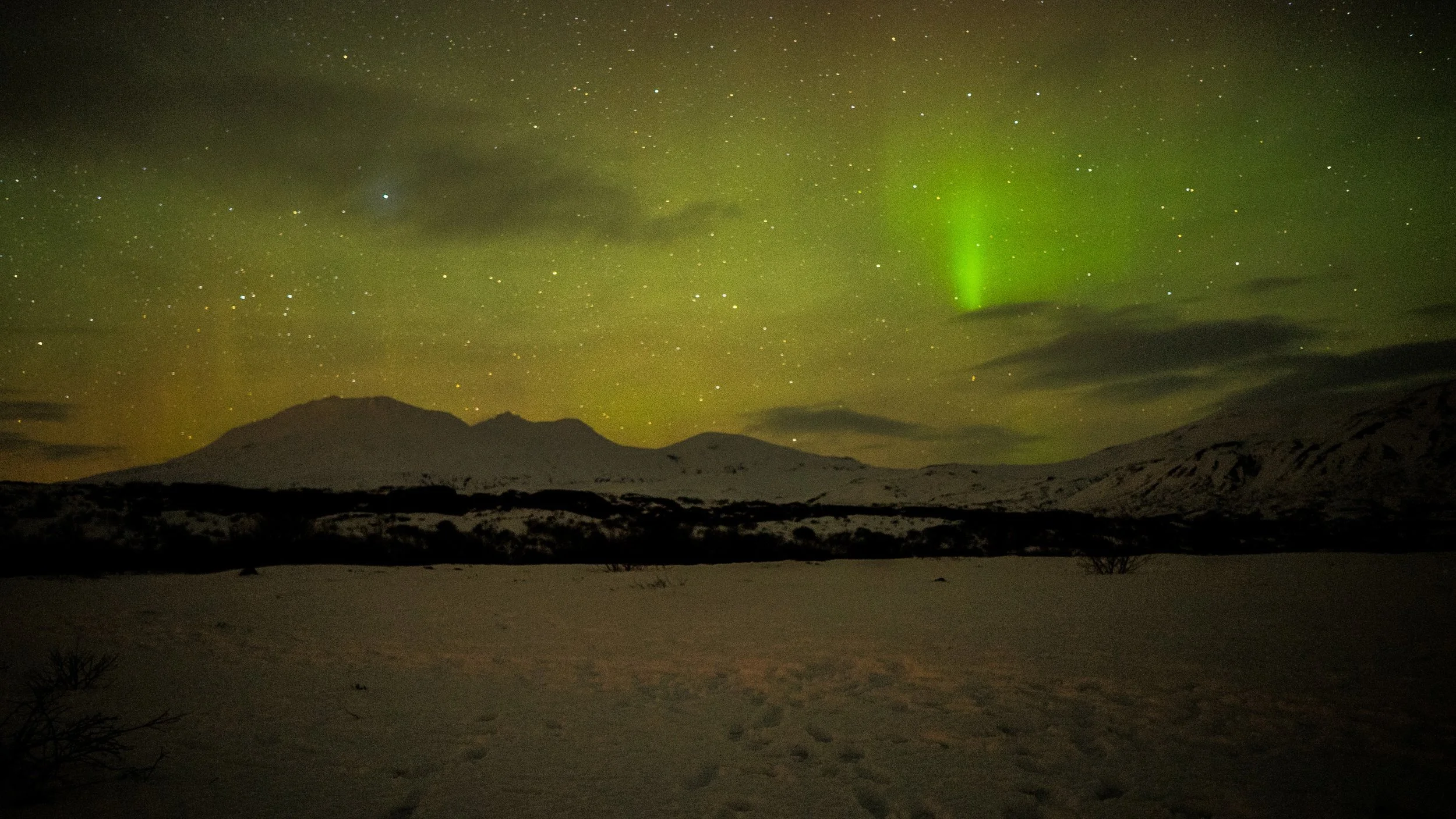 Snow-covered landscape with mountains under a starry sky and green aurora borealis lights.