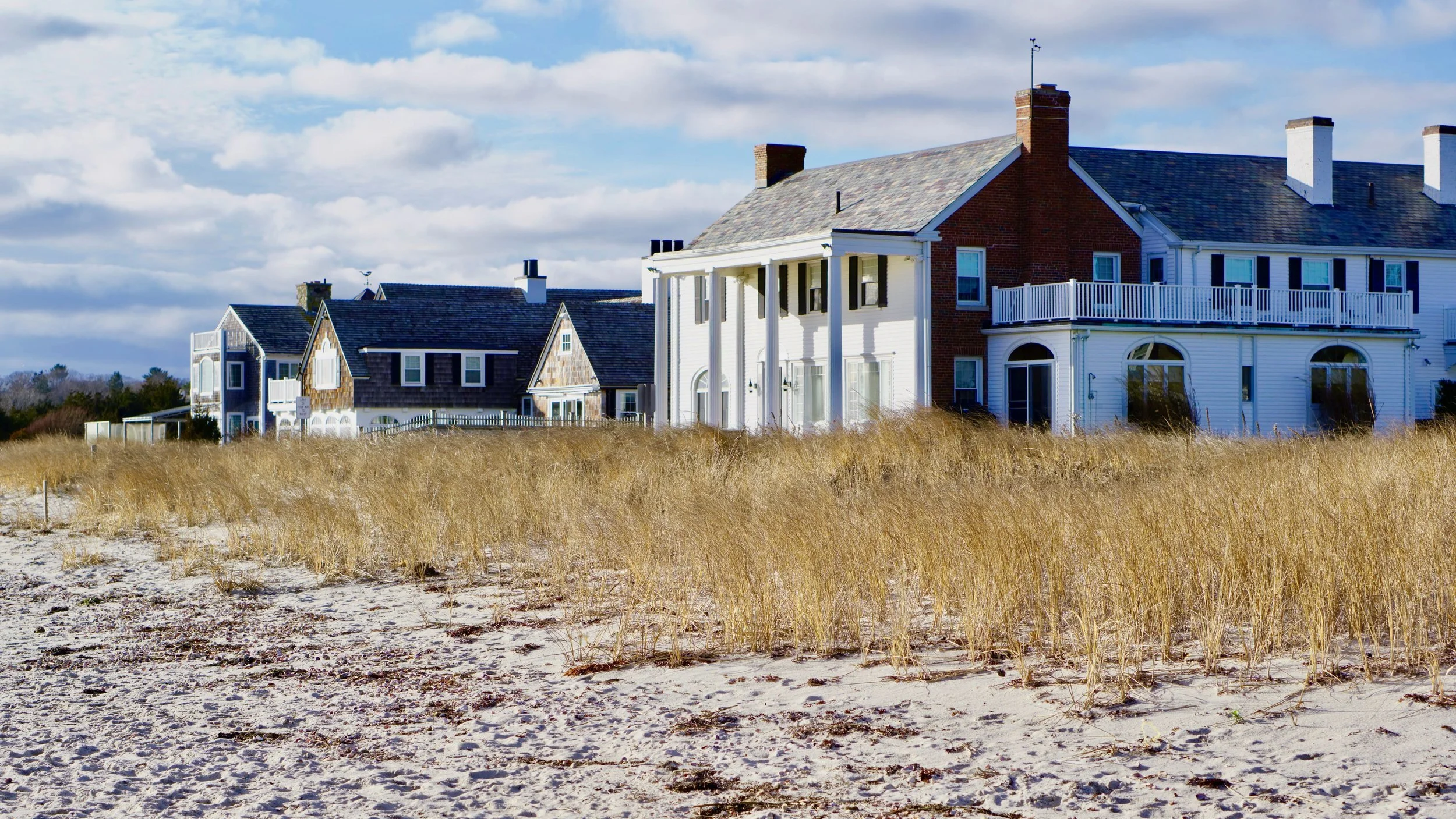Beachfront houses with grassy dunes and sandy beach under a partly cloudy sky.