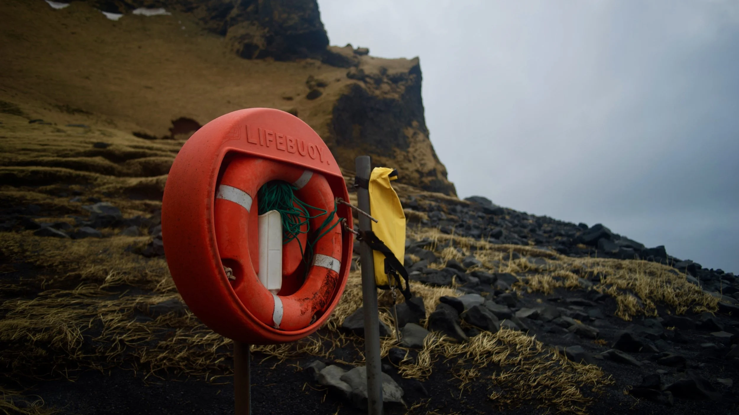 Orange lifebuoy with rope mounted on a post near a rocky, grassy hill.