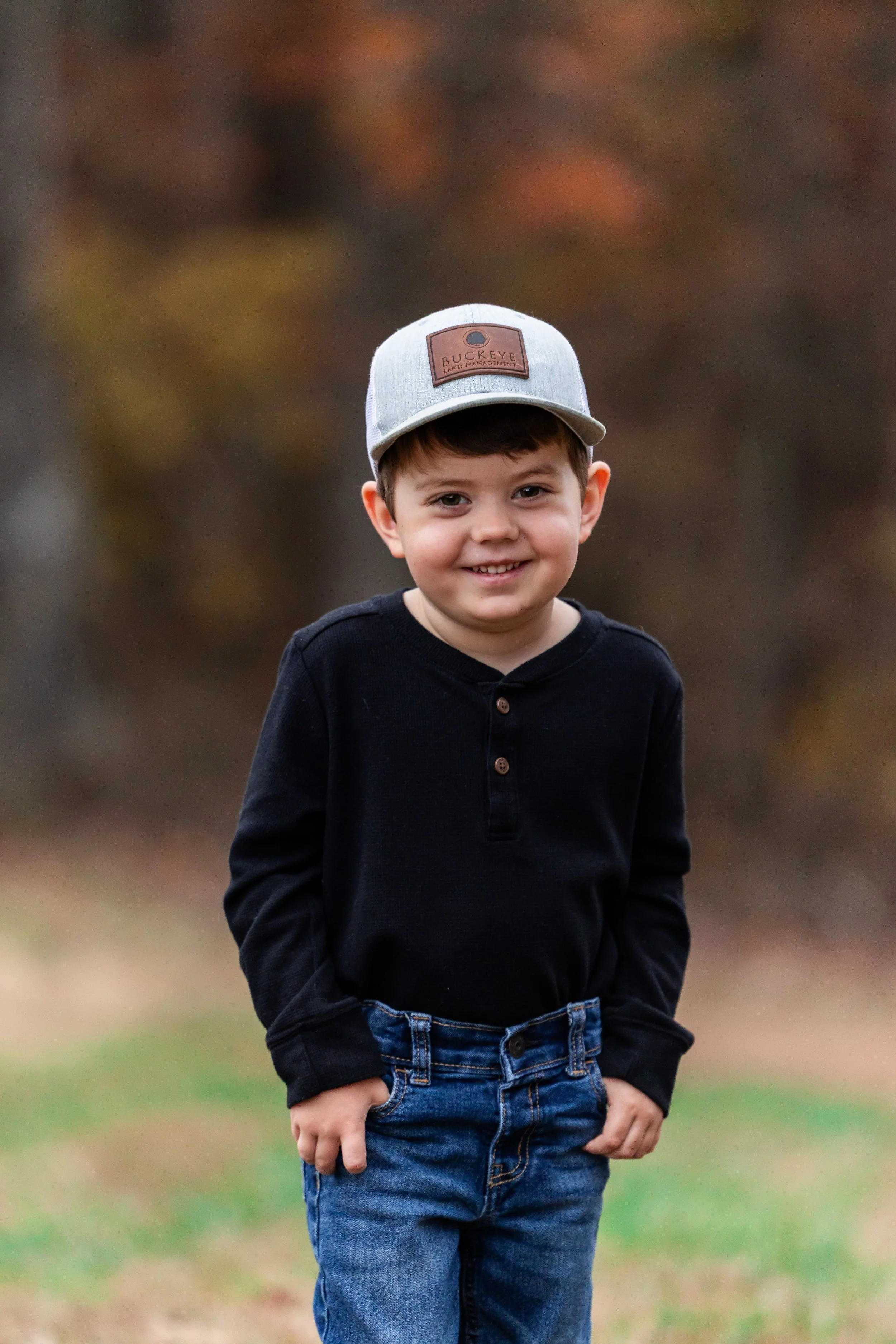 A young boy standing outdoors on a cloudy day, wearing a gray and brown baseball cap with a logo, a black long-sleeve shirt, and blue jeans, smiling at the camera.