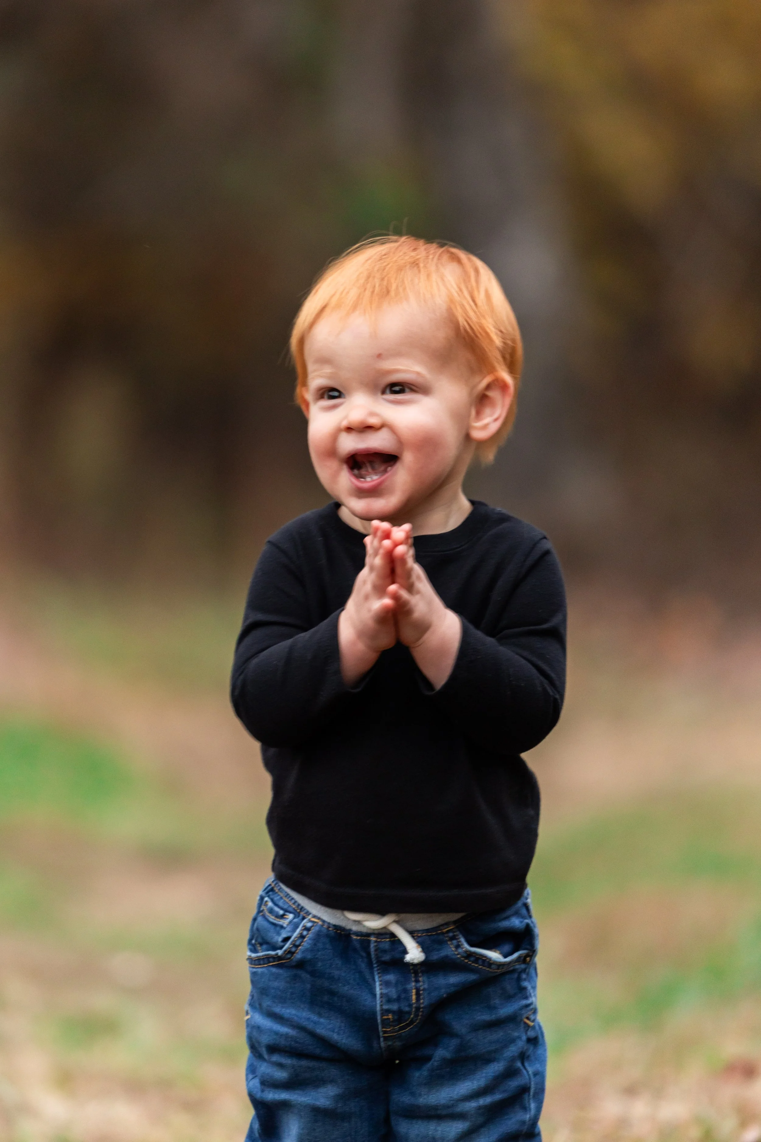 A young boy with red hair and a big smile, wearing a black long-sleeve shirt and blue jeans, standing outdoors with his hands pressed together in a gesture of happiness or excitement.