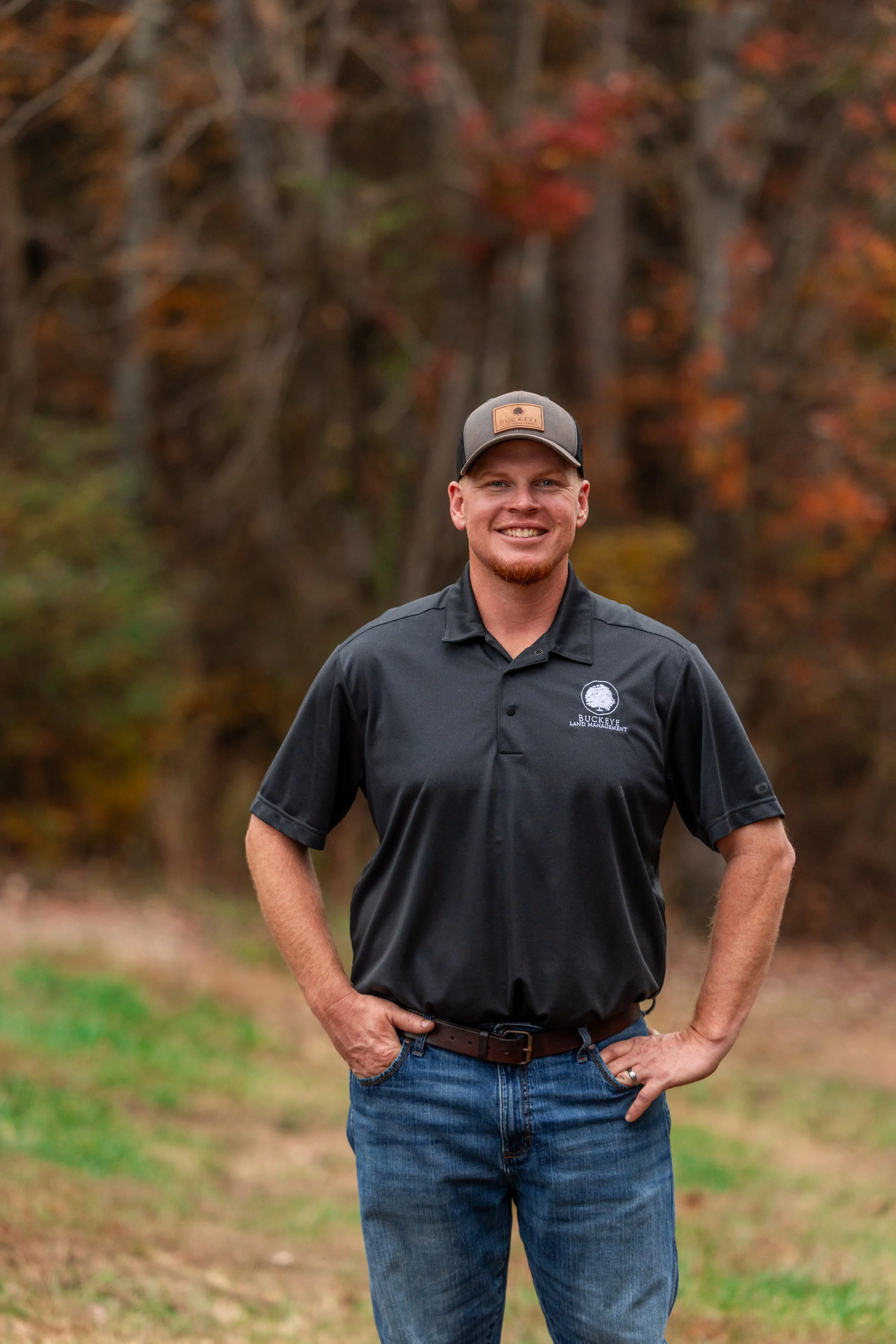 Smiling man in black polo shirt and jeans stands outdoors with trees showing fall colors in the background.