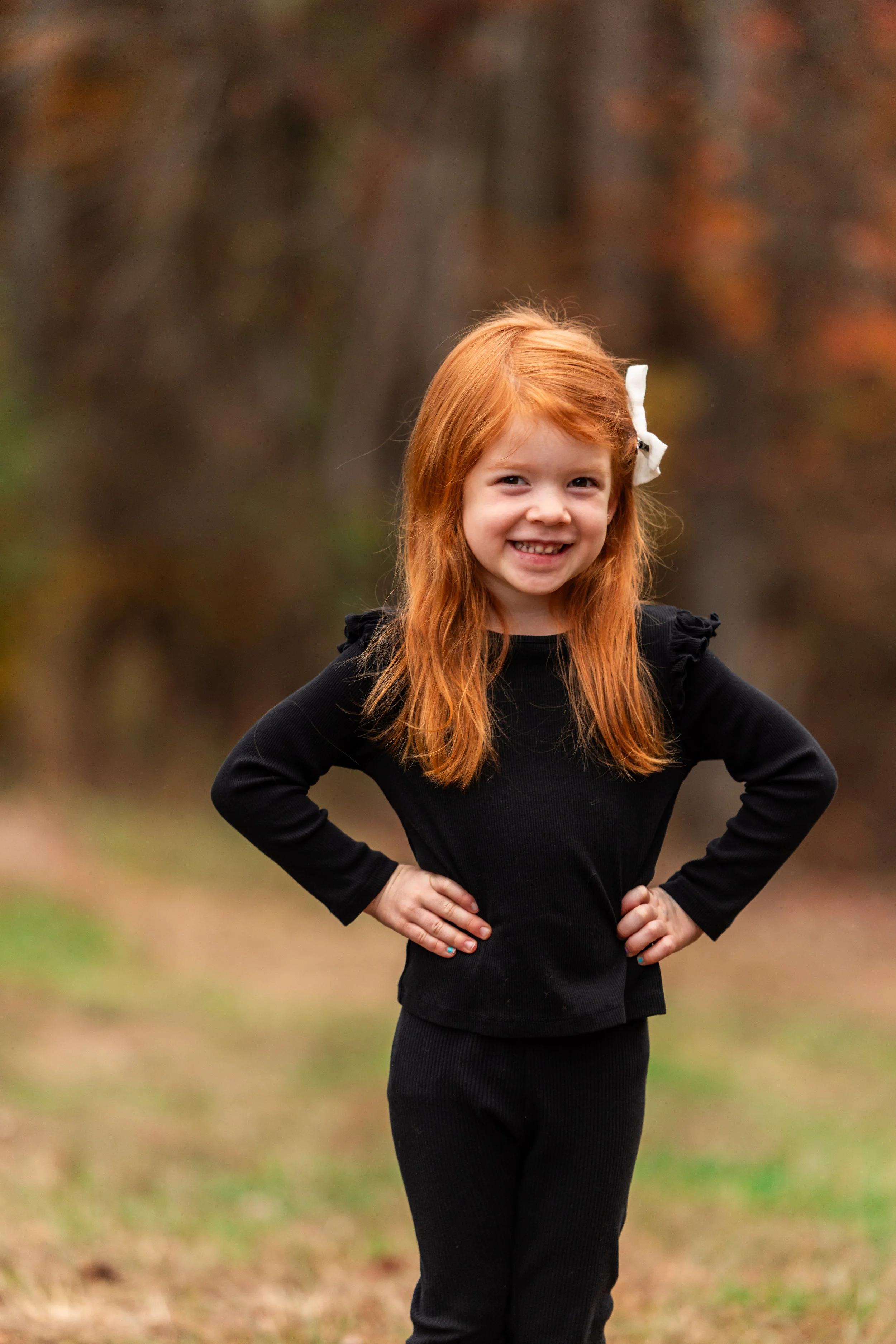 A young girl with red hair and a white bow, smiling, standing outdoors with her hands on her hips, wearing a black outfit, autumn trees in the background.