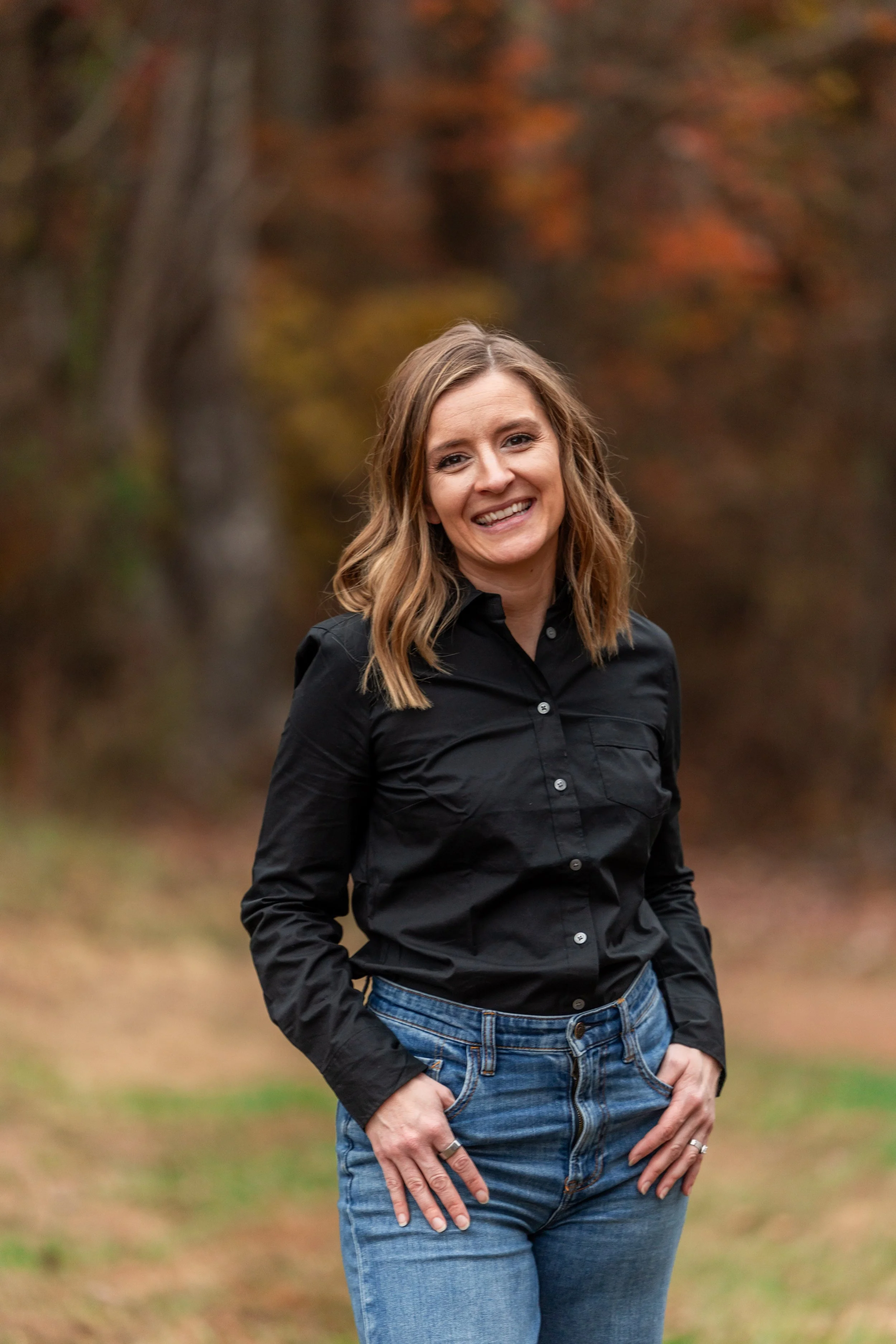 A smiling woman with shoulder-length light brown hair, wearing a black button-up shirt and blue jeans, standing outdoors in an autumn setting with blurred trees and fall foliage in the background.