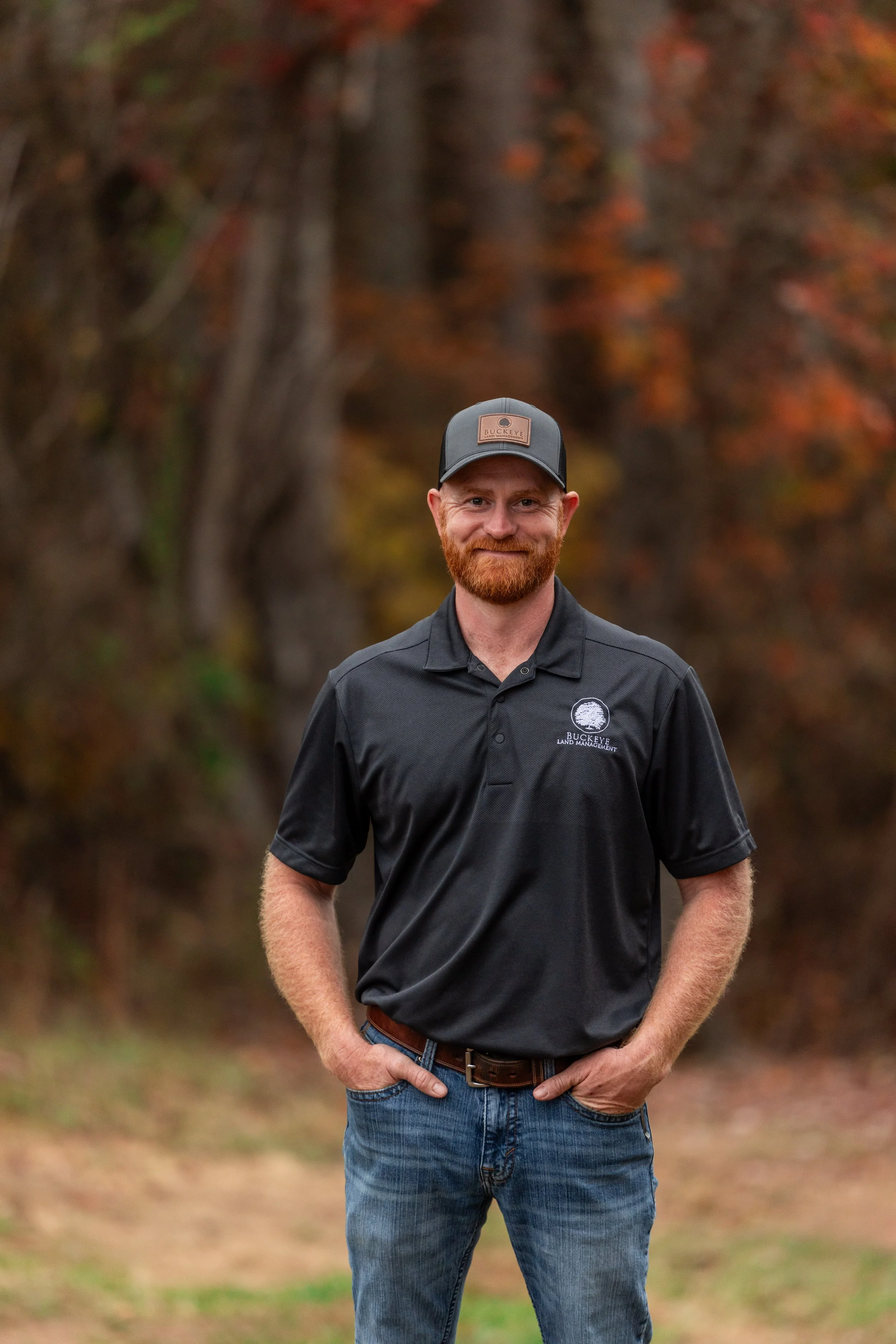 A man with a red beard and wearing a black polo shirt and baseball cap stands outdoors with autumn trees in the background.