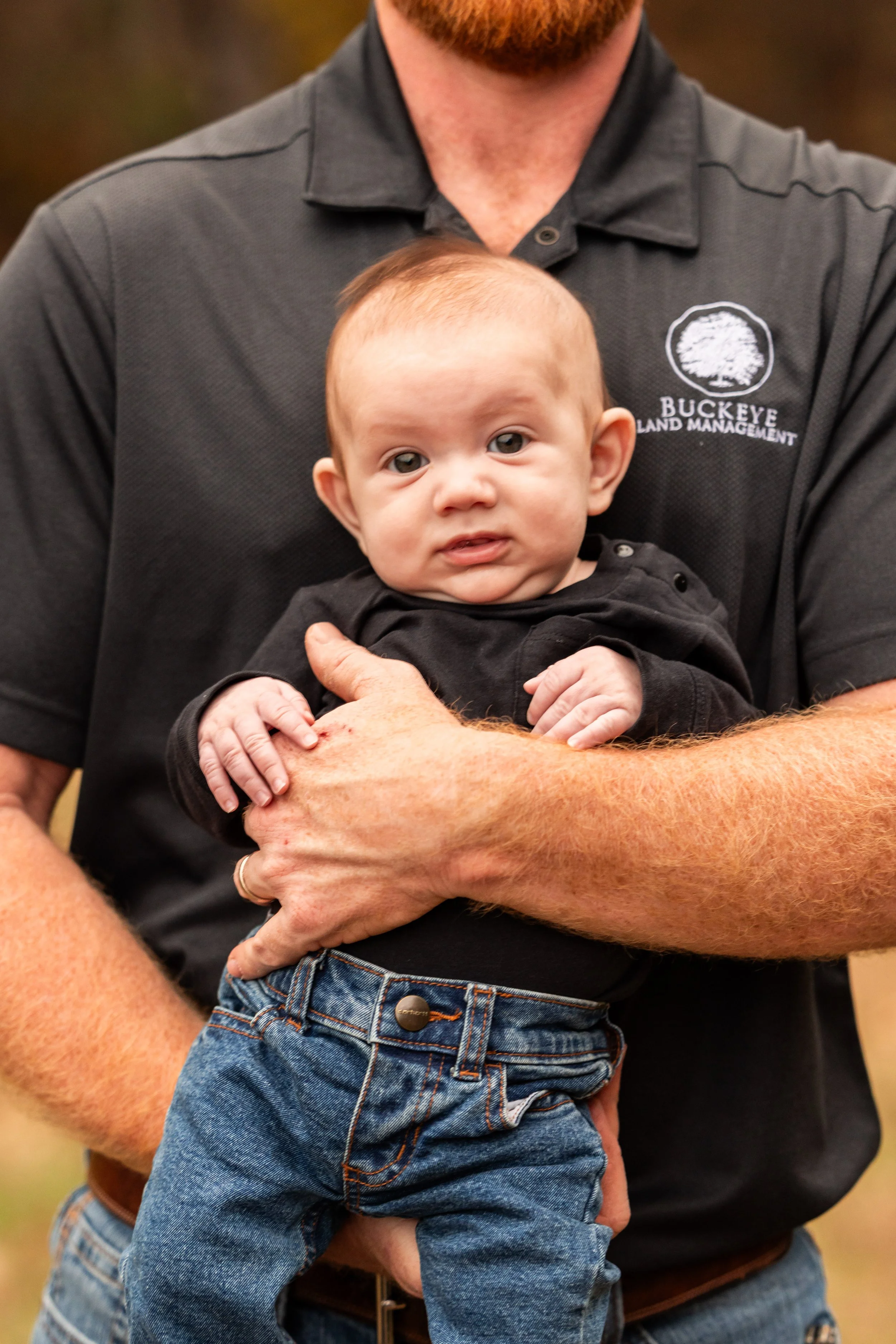 A man with a beard holding a baby with short light brown hair and blue eyes. The man is wearing a dark gray polo shirt with a logo that says Buckeye Land Management. The baby is dressed in a black shirt and blue jeans.