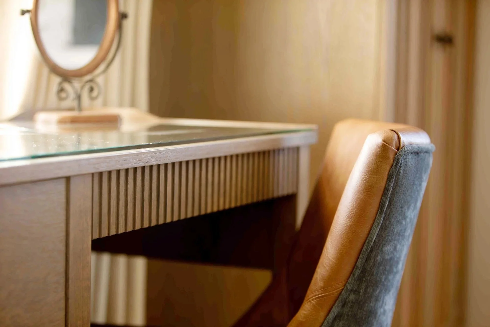 Close-up of a wooden vanity with a glass top and a brown and gray upholstered chair in a warmly lit room.
