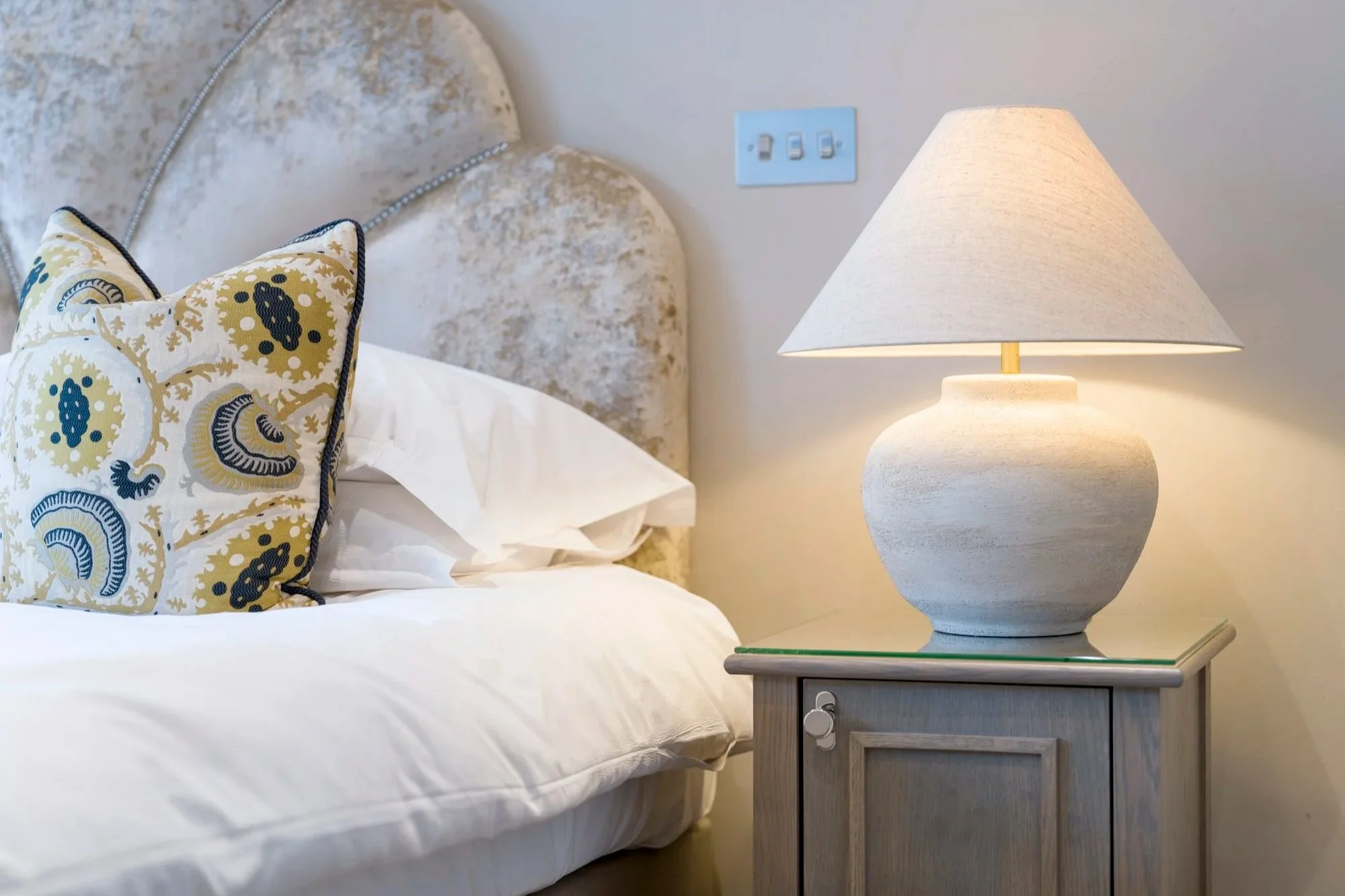 A bedroom bedside table with a white textured lamp, a decorative pillow with a floral pattern, a bed with white sheets, and a textured headboard with a wall switch in the background.