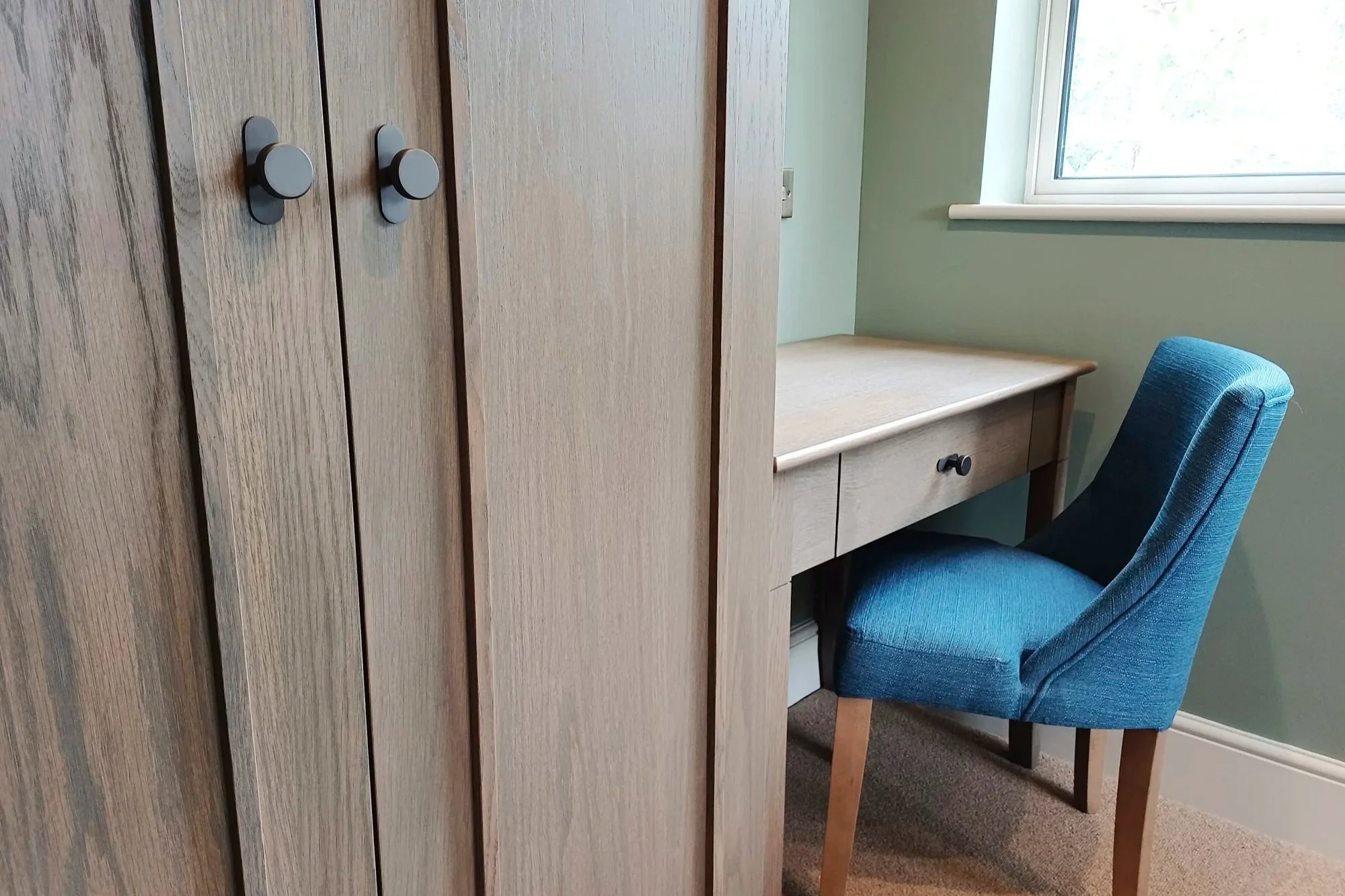 A cozy corner with a wooden wardrobe, a desk with drawers, a blue upholstered chair, and a window providing natural light.