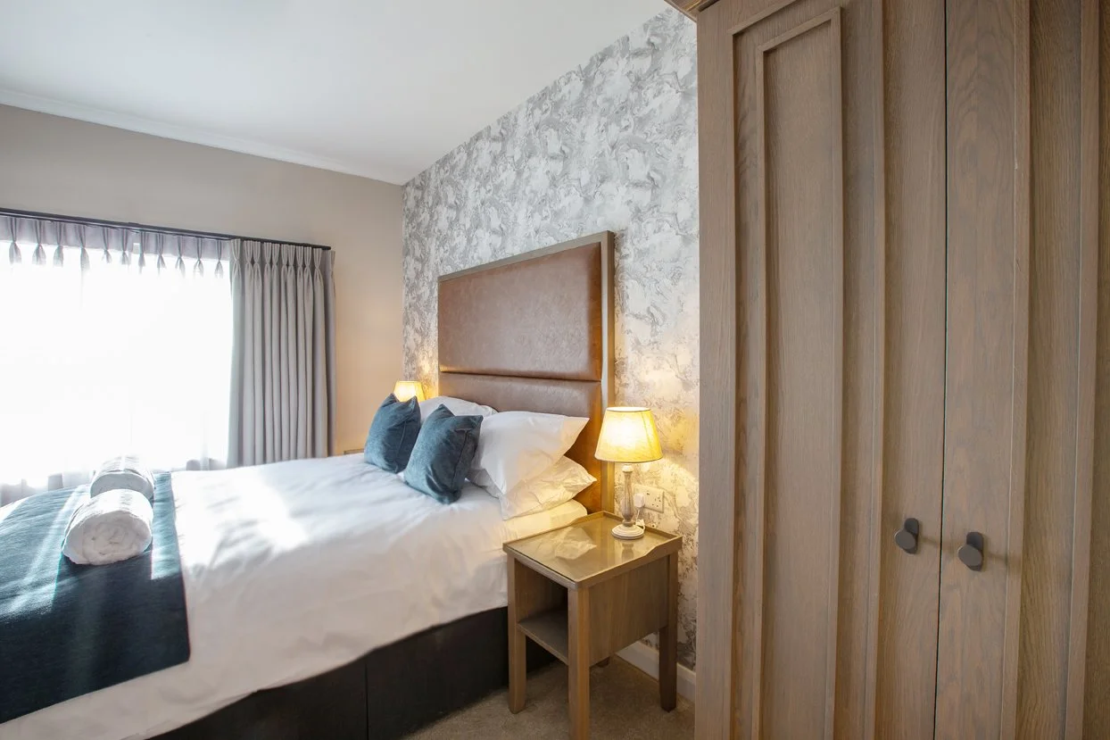 A cozy bedroom with a bed, pillows, bedside lamp, and a window with curtains. There is a wooden nightstand next to the bed and a wooden wardrobe.