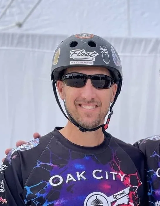 Person wearing a helmet and sunglasses, smiling, with an Oak City themed shirt.